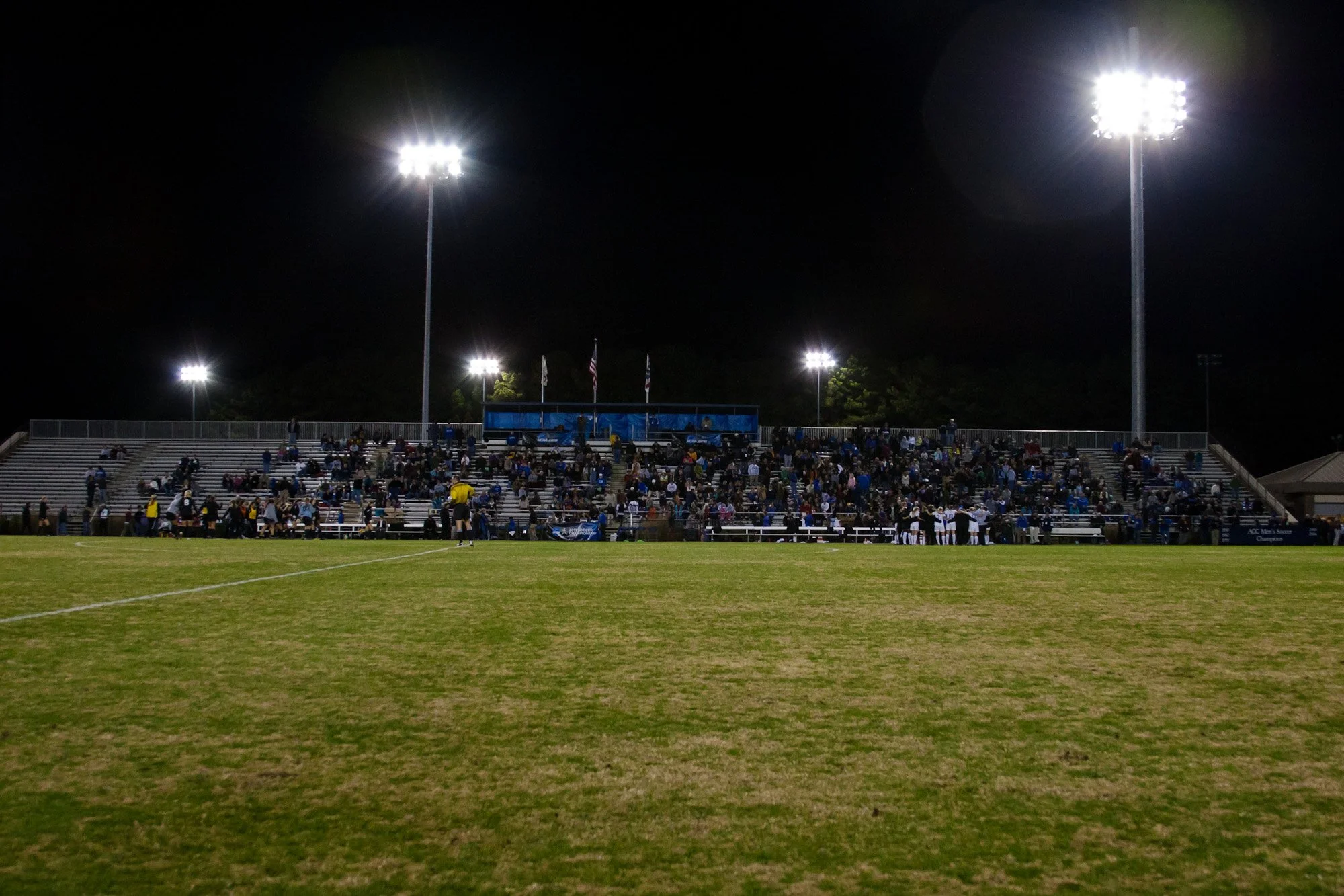 Soccer field at night illuminated by bright stadium lights, with spectators in the stands and players on the field.