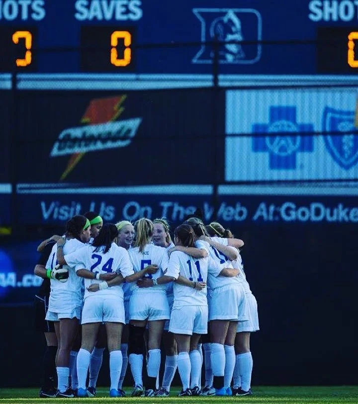 Group of female soccer players in white uniforms huddling together on the field before a game, with a scoreboard and advertisement screens in the background.