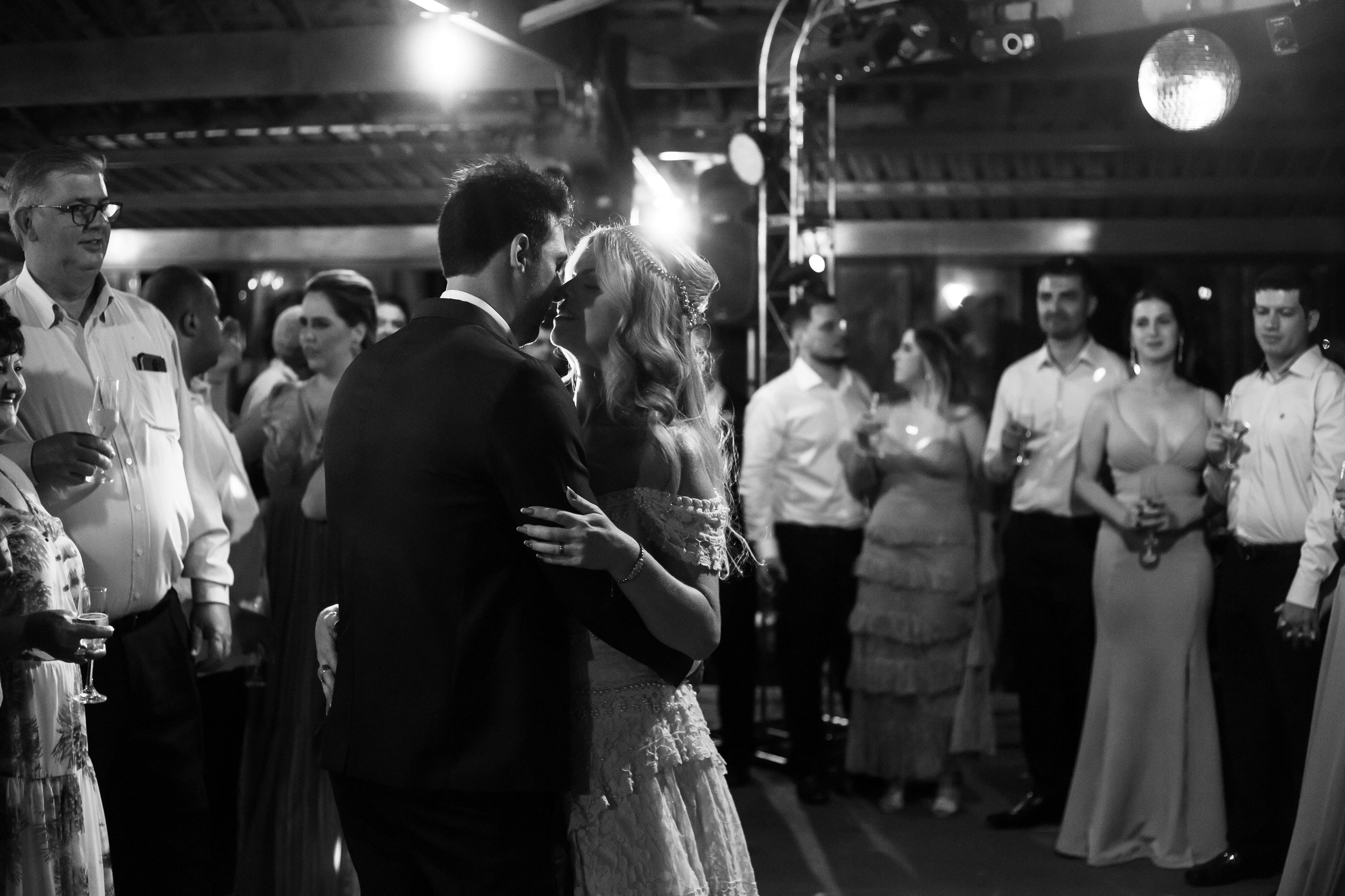 A black-and-white photo of a wedding reception where a bride and groom are dancing closely, surrounded by guests holding drinks and watching them.