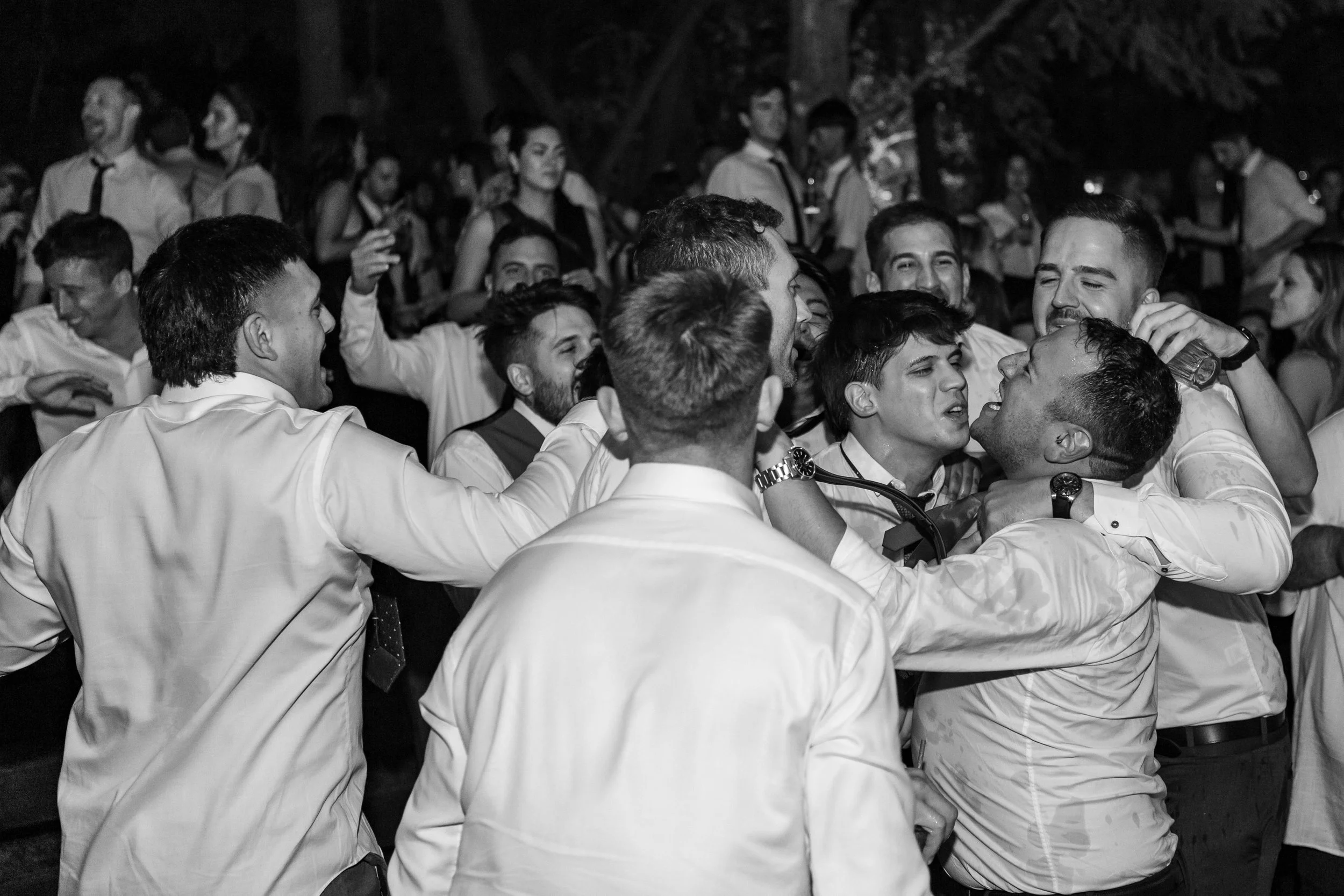 A group of men in white shirts and dark pants are engaged in a lively, playful fight or dance, with some tugging or holding each other, surrounded by other people at a social gathering or party.