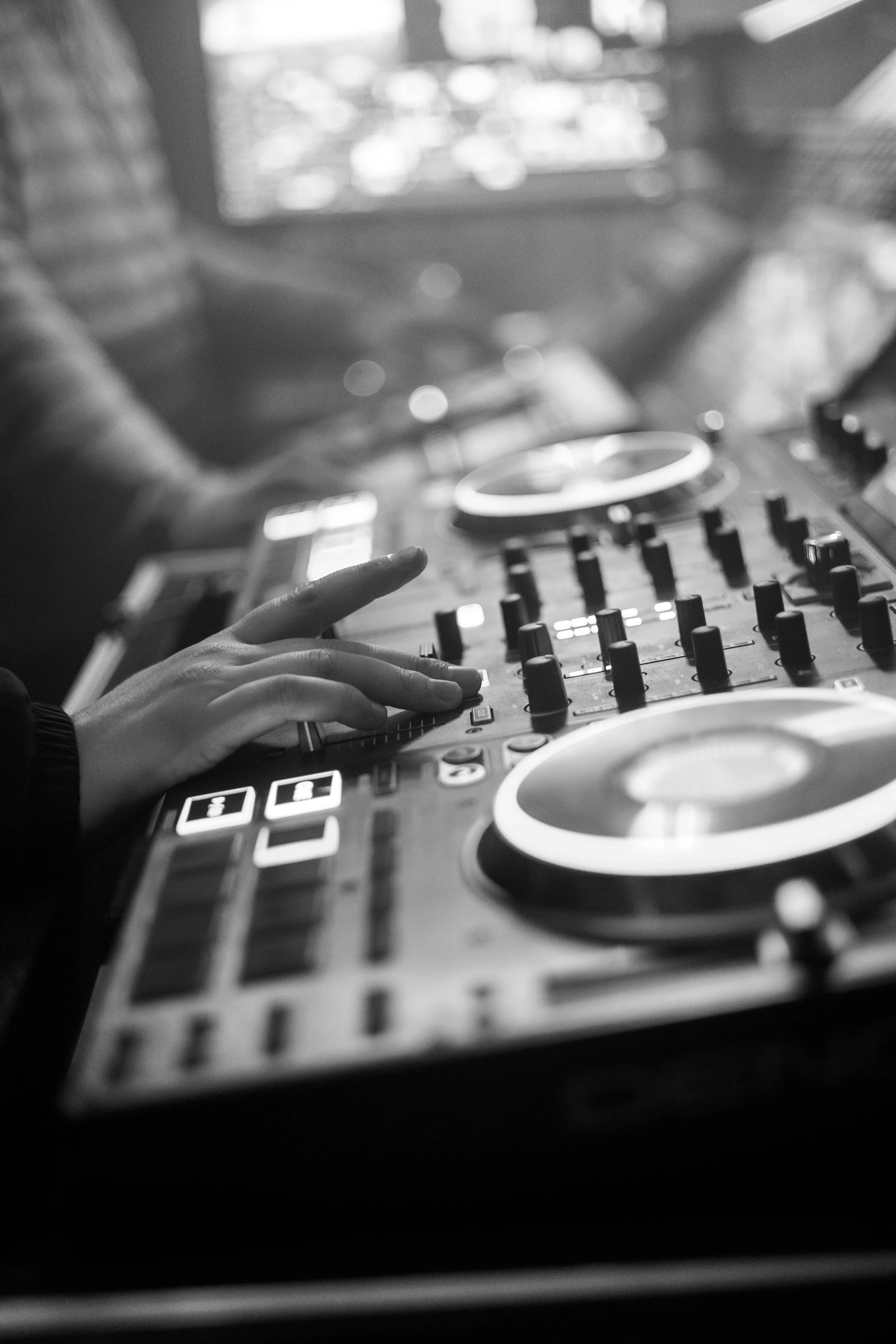 Close-up of a DJ's hand on a turntable, with other DJ equipment visible, in a black-and-white photo.