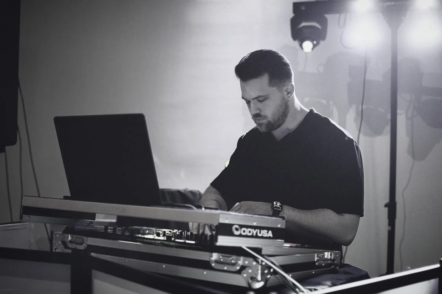 A man DJing with a laptop and DJ equipment in a dimly lit studio or club, black and white photo.