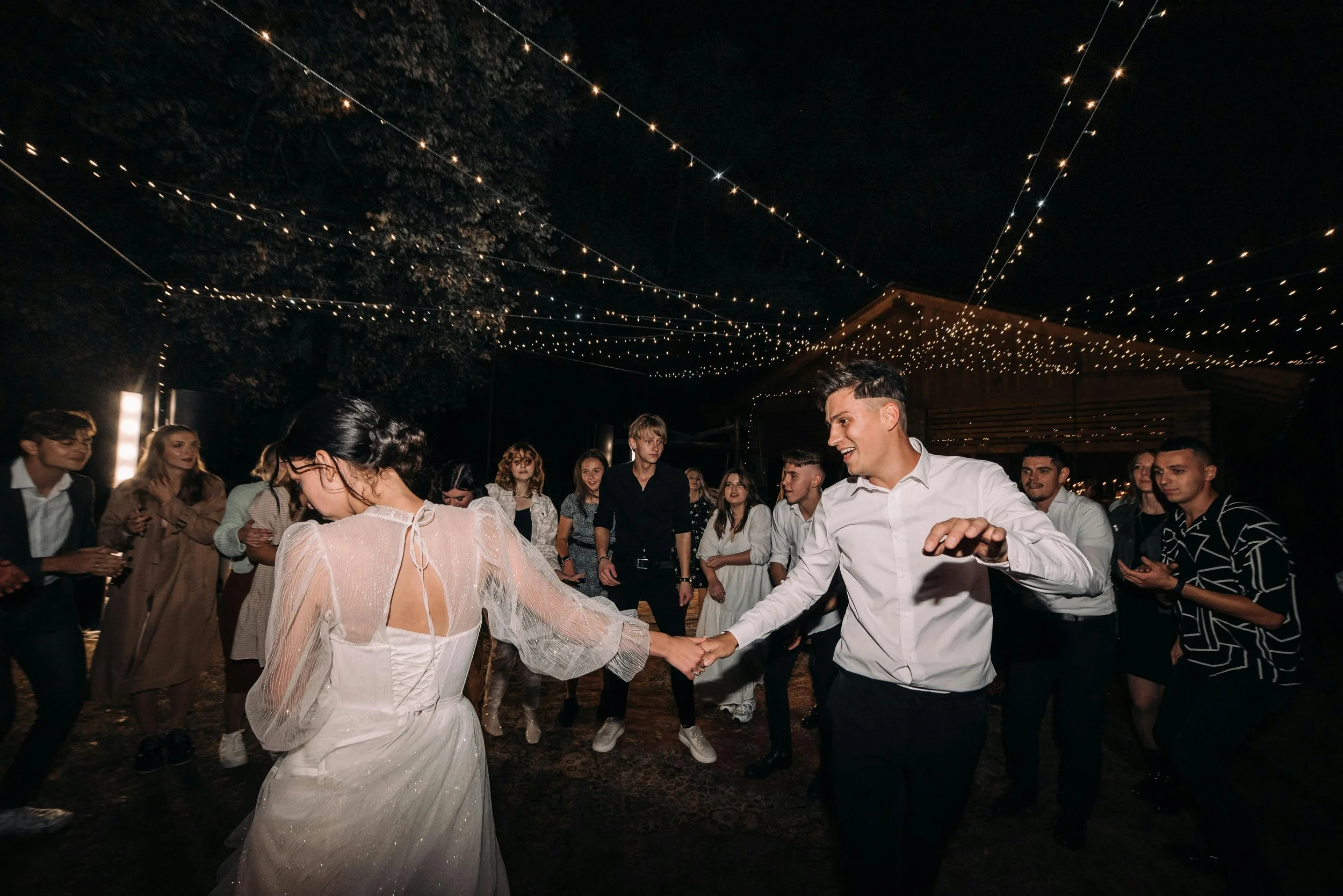 People dancing at a nighttime celebration under string lights, with a woman in a white dress and a man in a white shirt holding hands and smiling. Others are watching and enjoying the event.