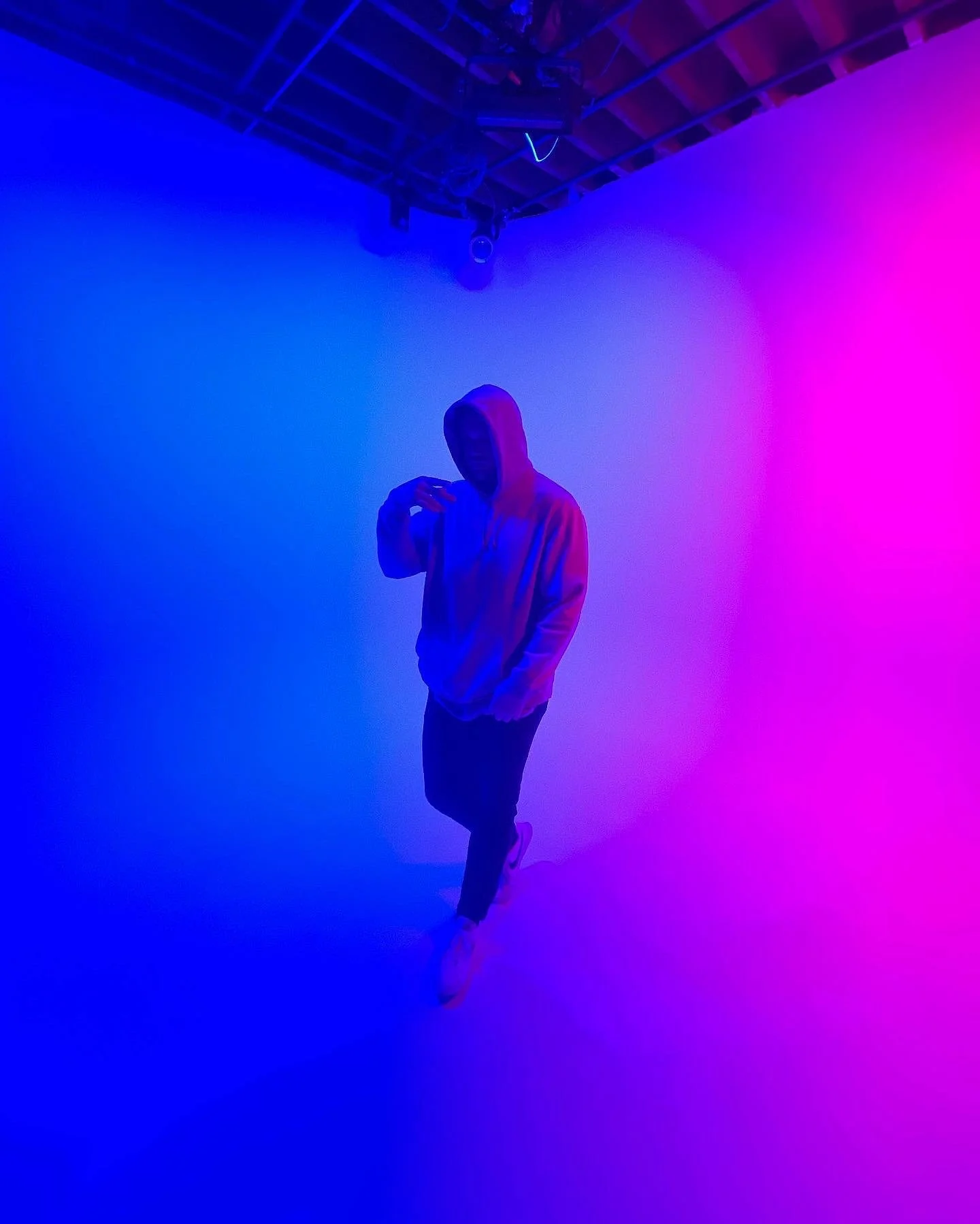 Person in a hoodie standing in an art studio with colorful pink and blue lighting.