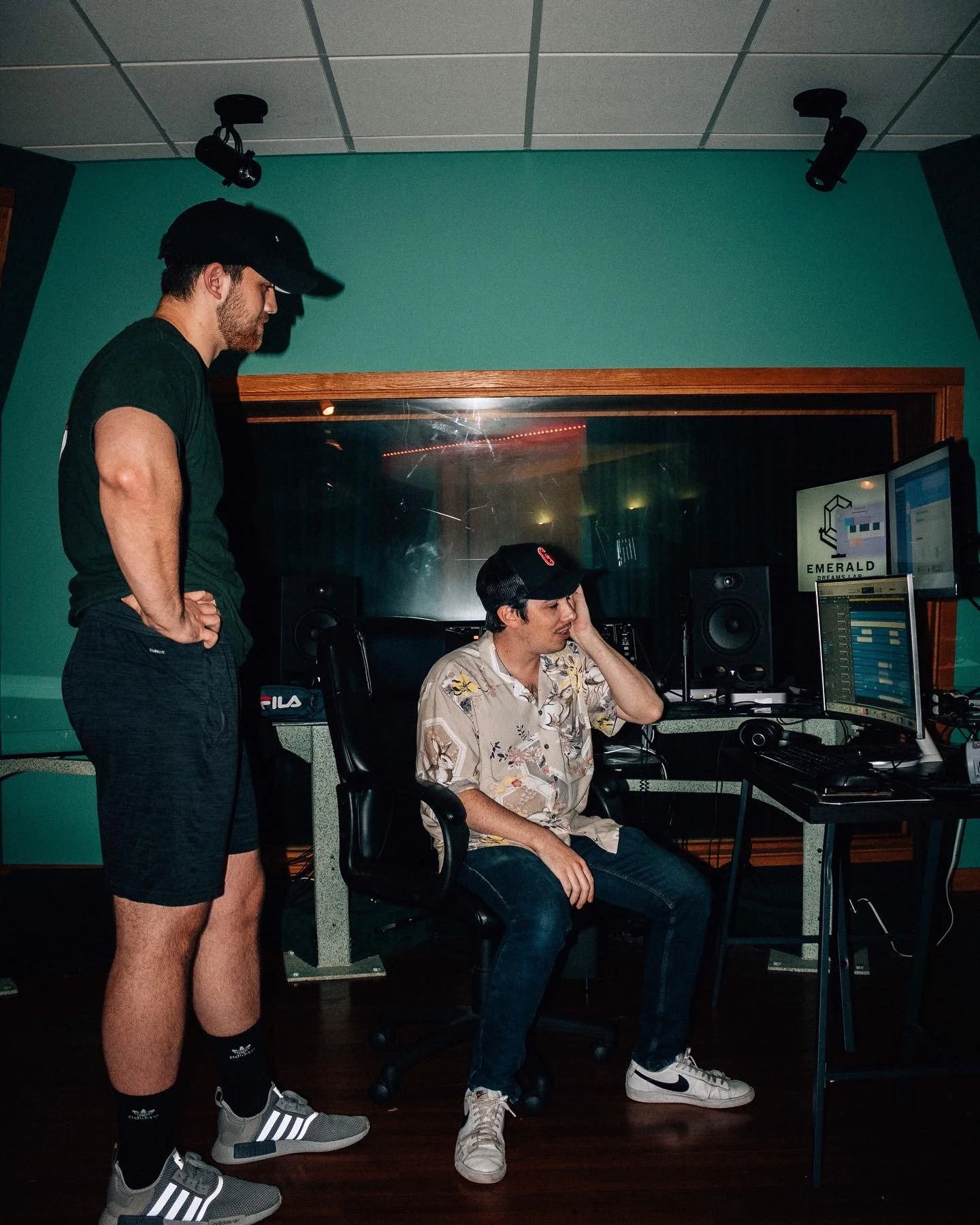 Two young men in a recording studio, one sitting at a desk with a computer and music equipment, the other standing nearby watching. Both are wearing casual clothes and baseball caps.