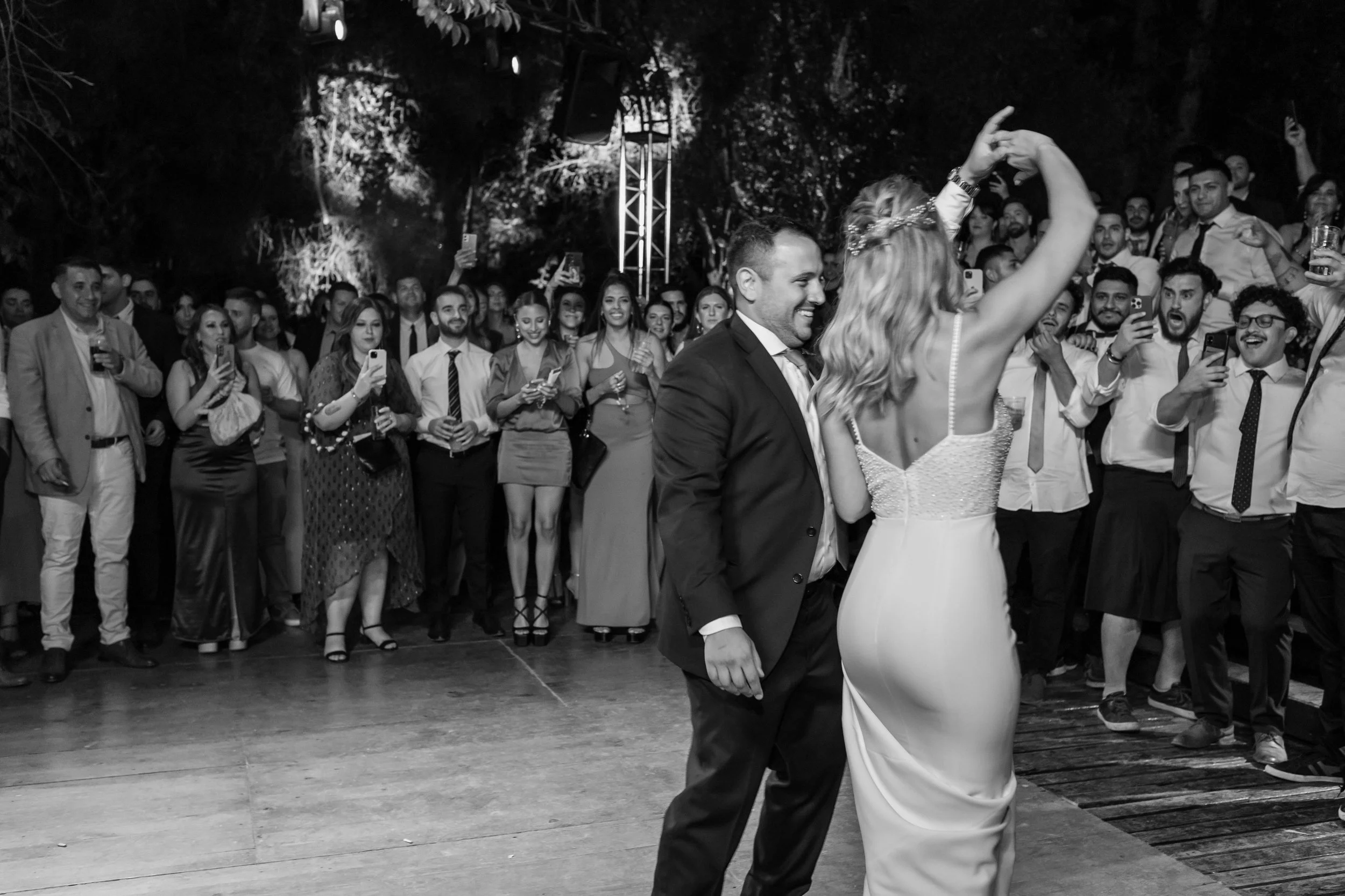 A black and white photo of a wedding reception with a bride and groom dancing in the center, surrounded by guests taking photos and watching.