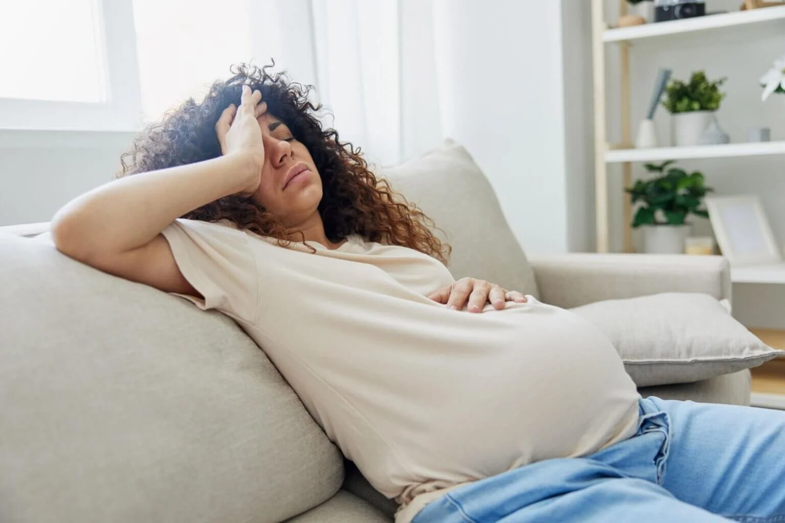 a pregnant woman is shown sitting on a couch with her hand on her forehead, appearing exhausted or fatigued.
