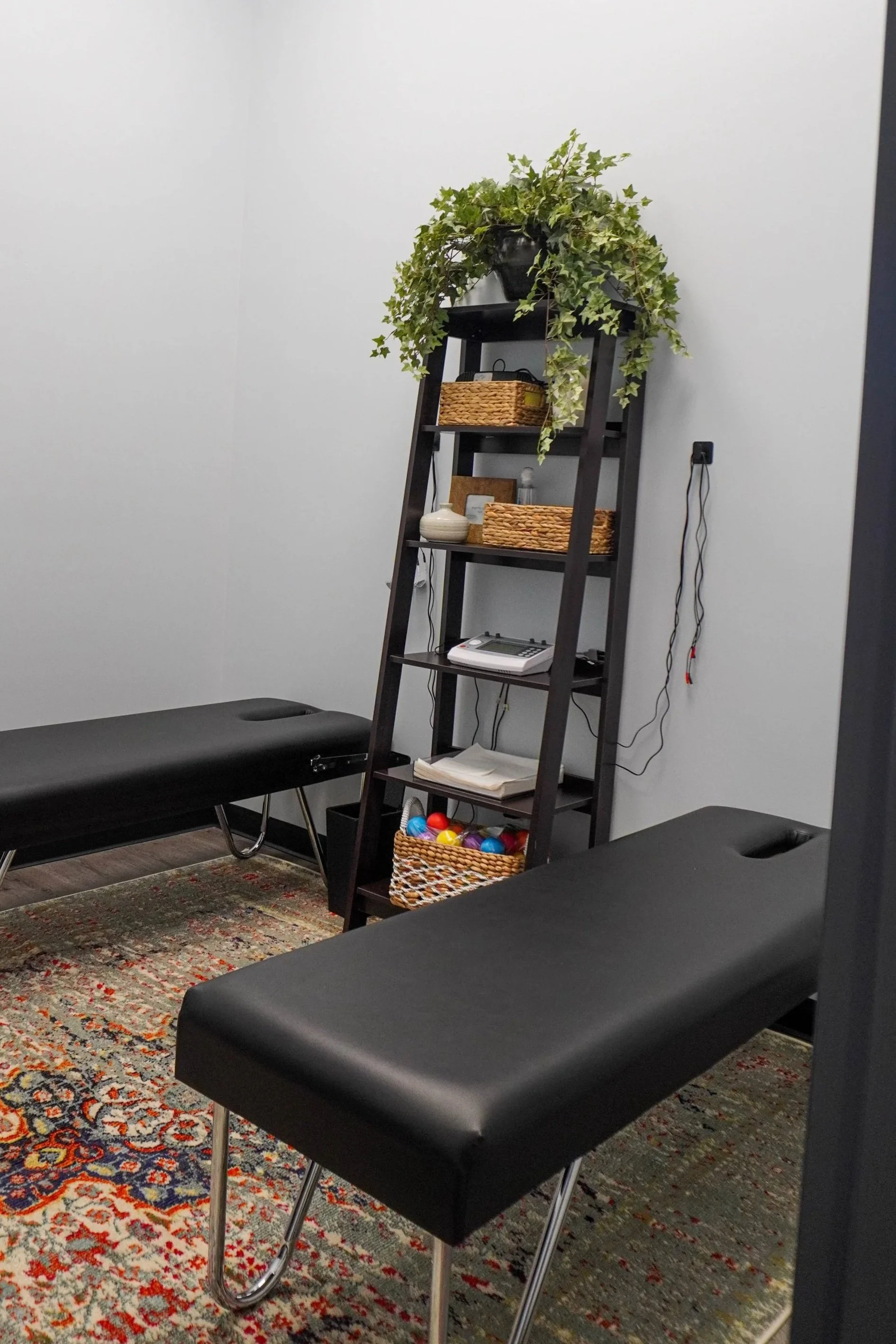 A room with two black therapy or massage tables, a tall black shelf with baskets, decorative items, a landline phone, and a basket of colorful plastic balls, a ceiling plant on top of the shelf, a patterned area rug, and white walls.