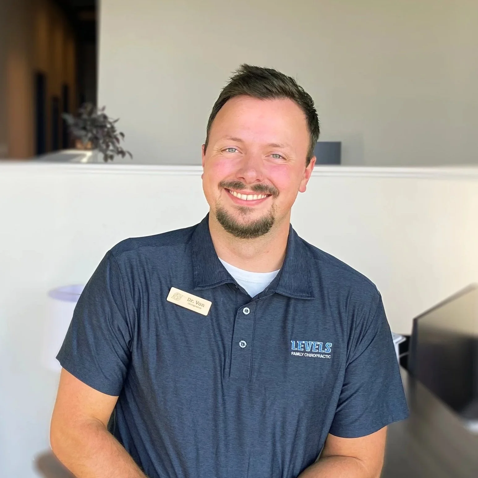 A smiling man with short dark hair and a beard, wearing a navy blue polo shirt with a name tag and the logo of LEVENS FAMILY CHIROPRACTIC, standing indoors with a blurred background.