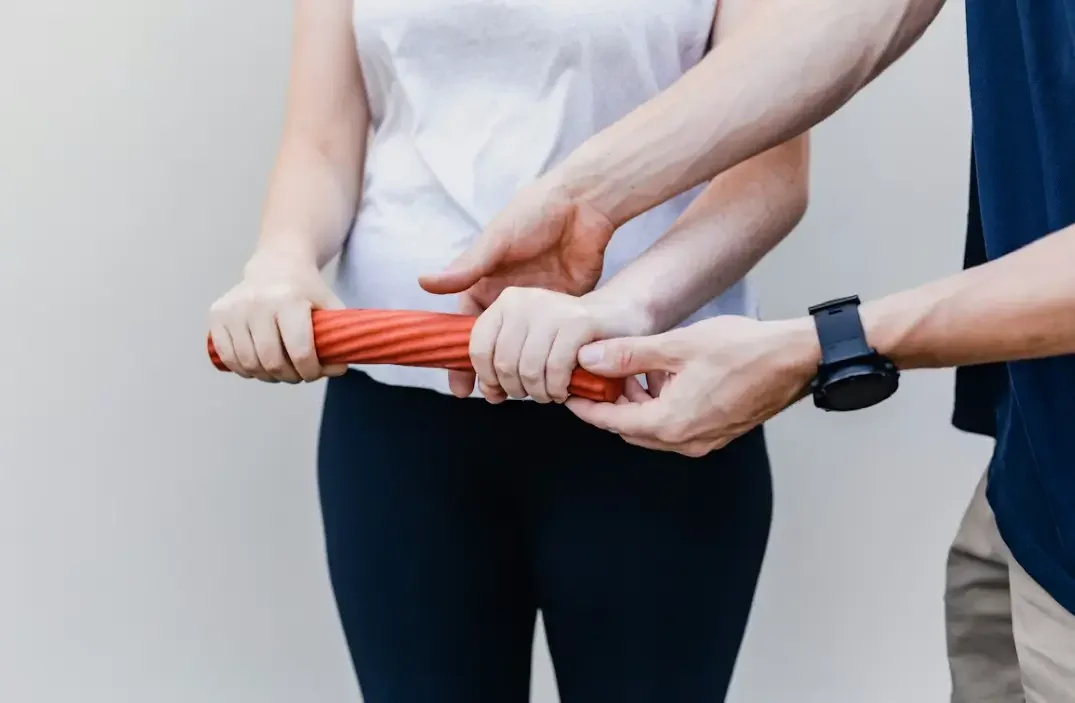 The image shows a physical therapist or professional guiding a person in using a resistance bar for rehabilitation exercises.