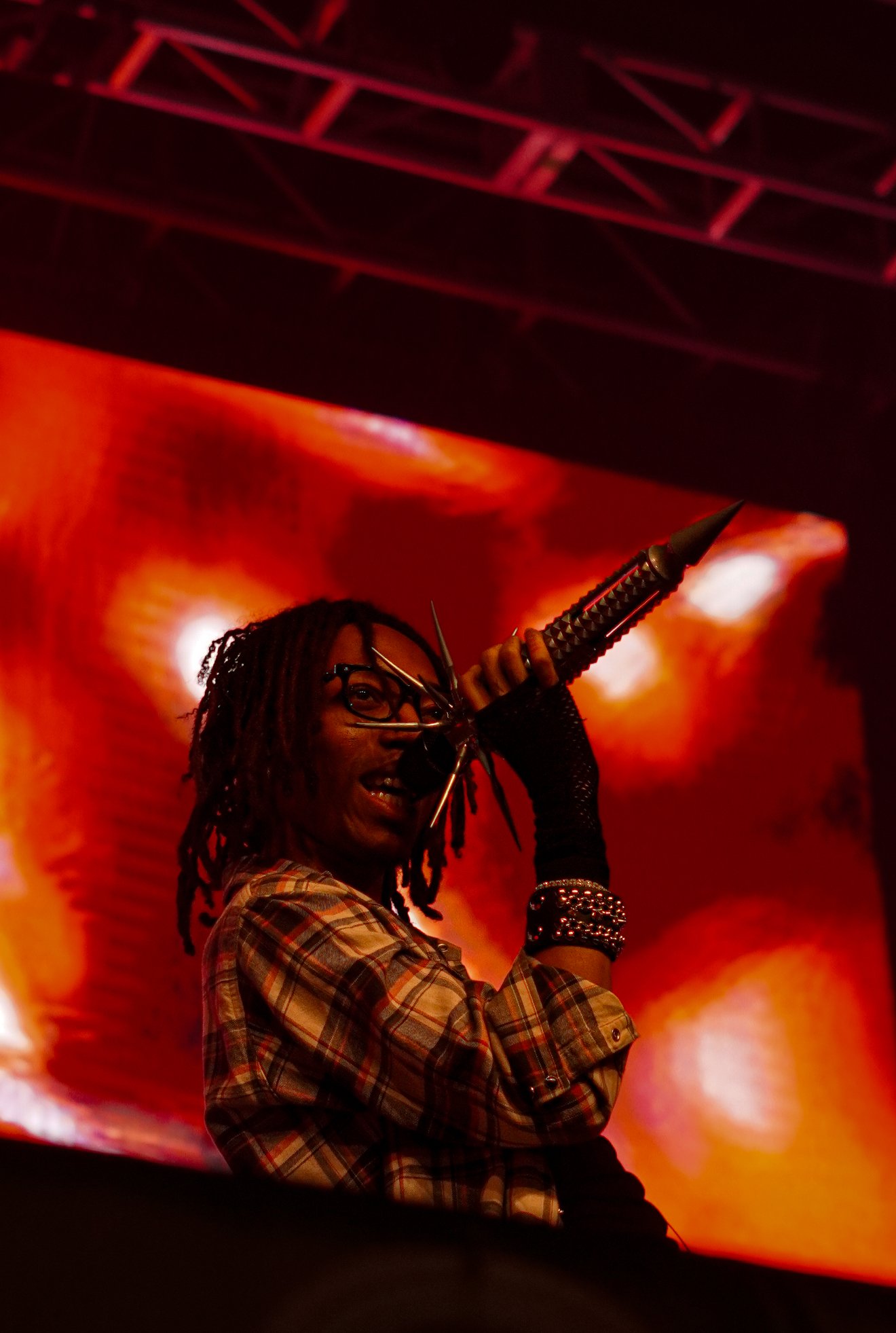 Performer with glasses and dreadlocks on stage, holding a microphone with a metal handle, singing or speaking, with a vibrant orange and red background.