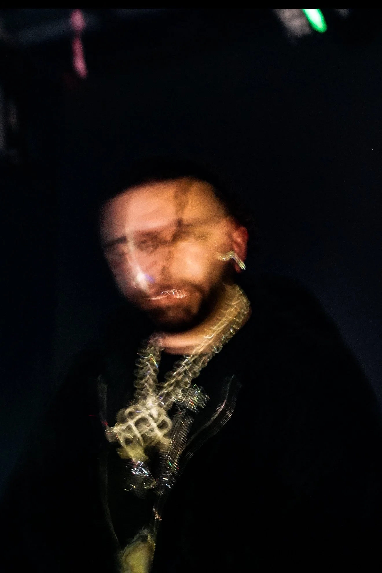 Blurry photo of a person with earrings and layered gold chains, in a dark setting with some neon lighting.