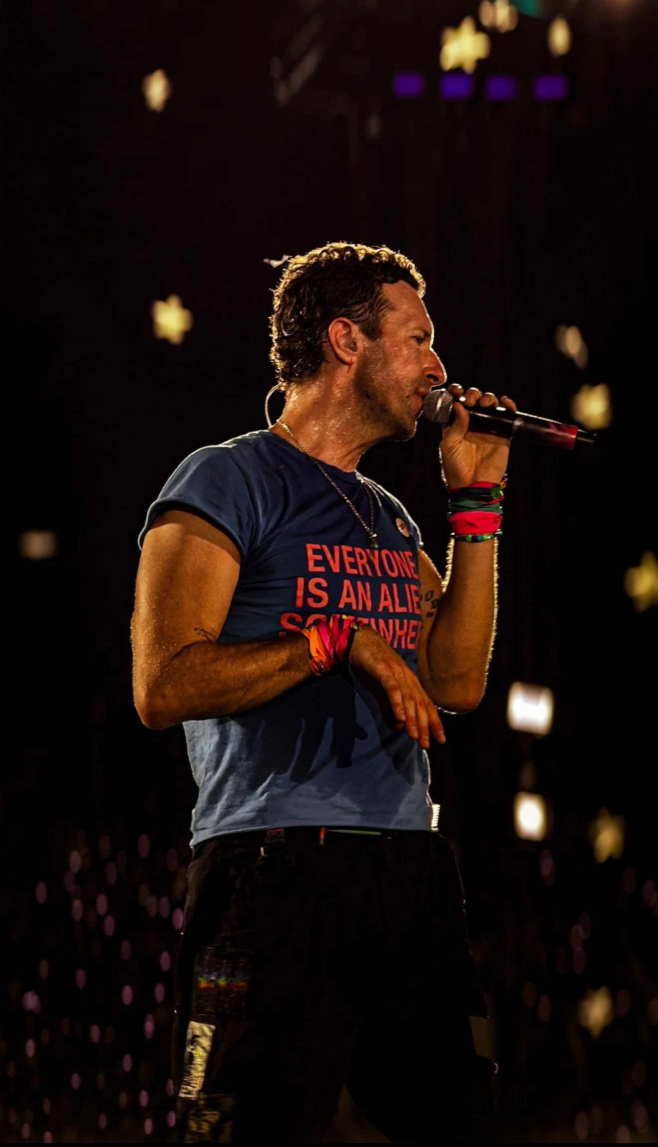 A male performer singing on stage, holding a microphone, wearing a blue T-shirt with red text, and colorful wristbands, with a dark background and star-shaped lights.