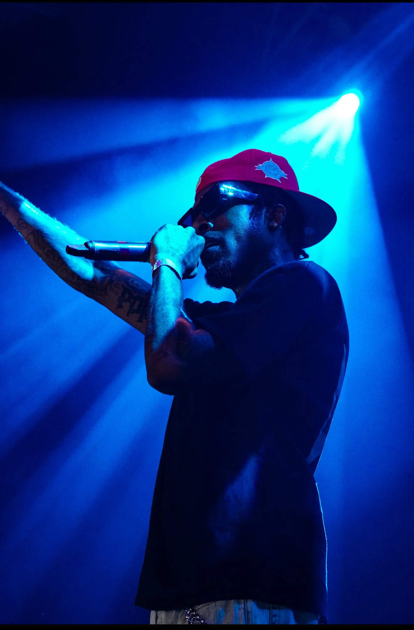 A male performer wearing a red cap, sunglasses, and a black T-shirt is holding a microphone and performing on stage illuminated by blue stage lights.