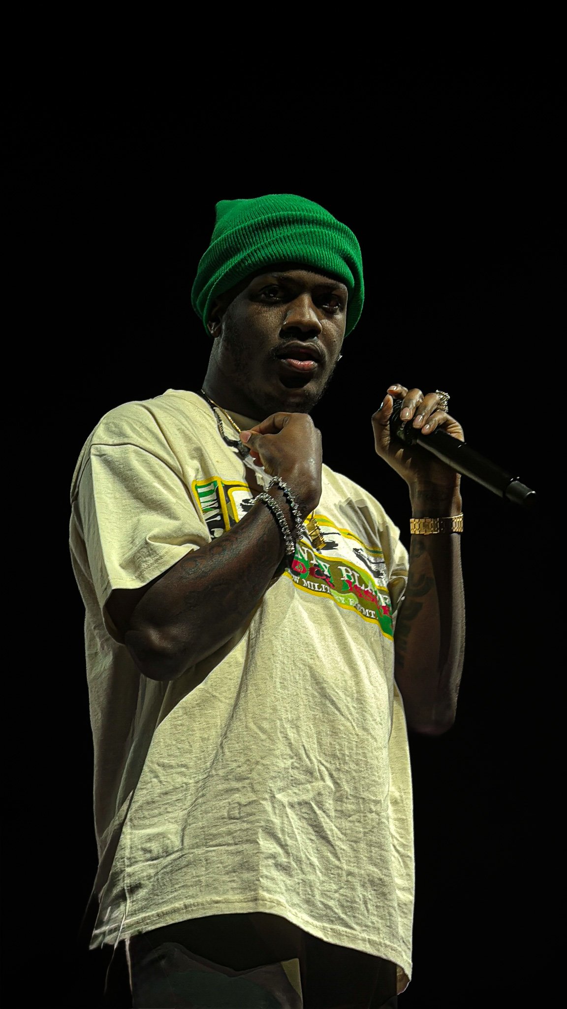A man wearing a green beanie, a beige T-shirt, and jewelry, holding a microphone on stage against a dark background.