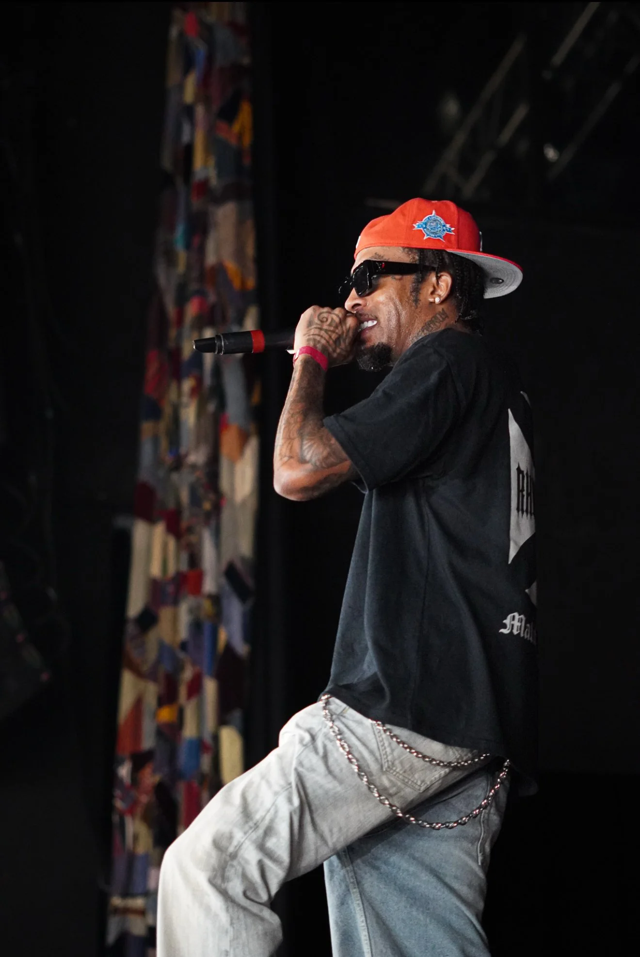 A male rapper performing on stage, wearing a red baseball cap, black sunglasses, a black T-shirt, and light jeans with a chain. The background is dark with some colored fabric panels.