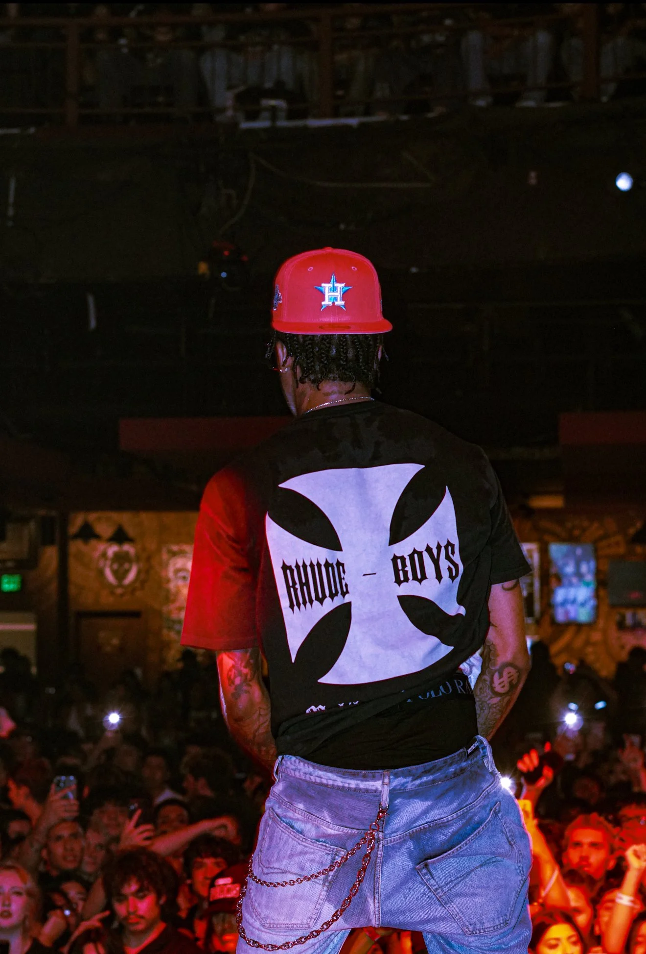 A performer on stage at a concert, facing away from the camera, wearing a red baseball cap and a black T-shirt with a bold design and text, with a crowd of fans in the background.