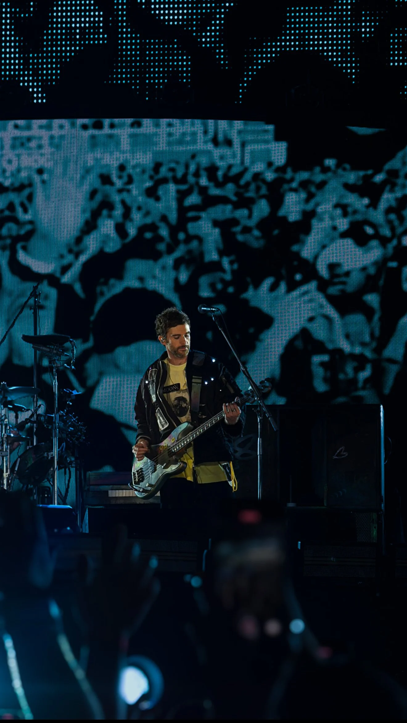 A musician playing bass guitar on stage during a concert, with a large digital backdrop displaying abstract patterns in blue and black.