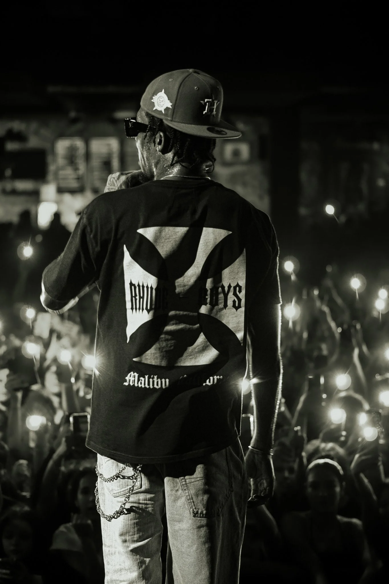 A performer on stage wearing a crowned cap and a T-shirt with the logo for Rhude Boys and Malibu King, facing away from the camera, with an audience holding up lights in the background.