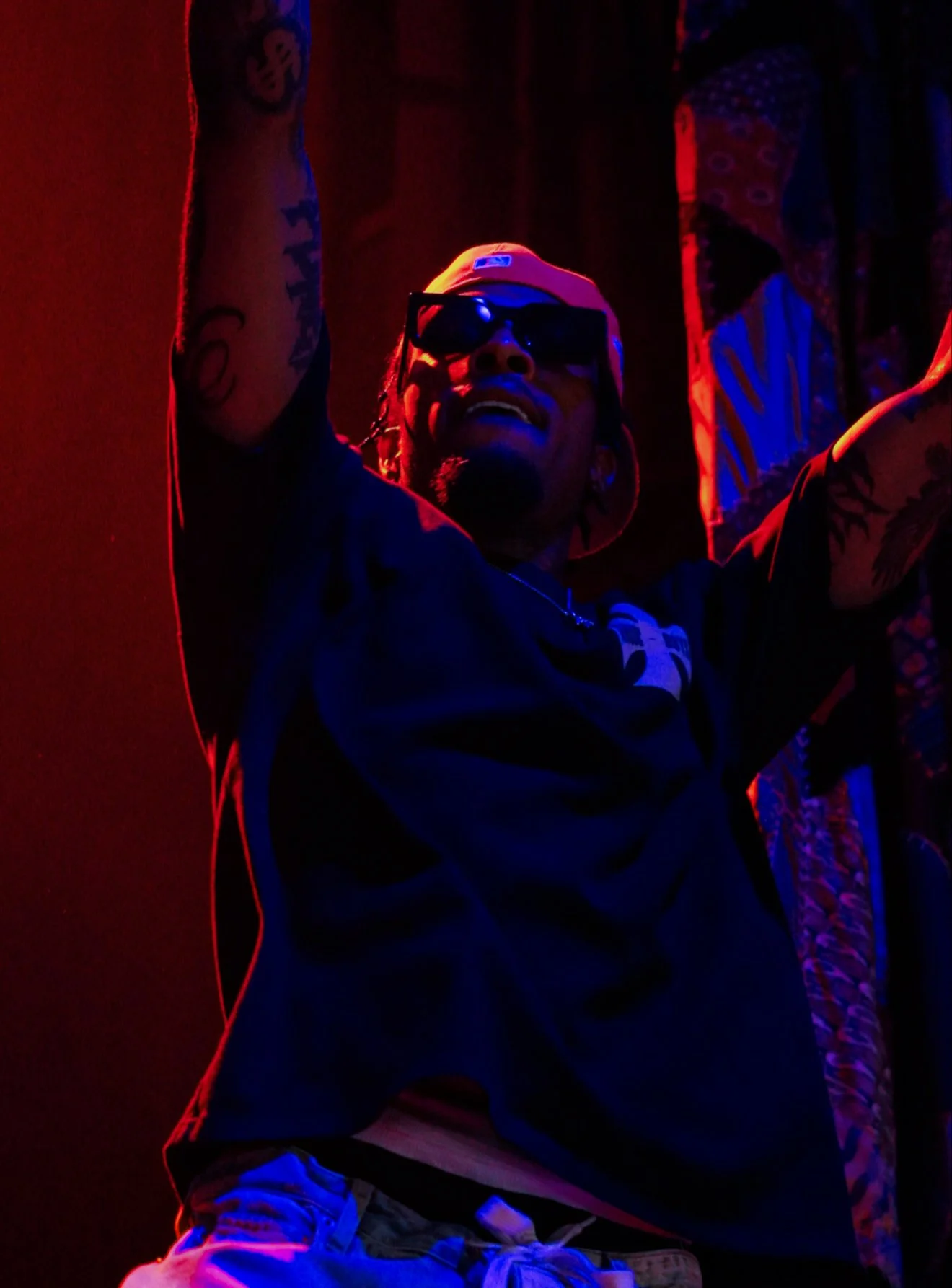 A man wearing sunglasses, a cap backwards, and a black shirt smiling with his arms raised in a dark, colorful setting with red and blue lighting.