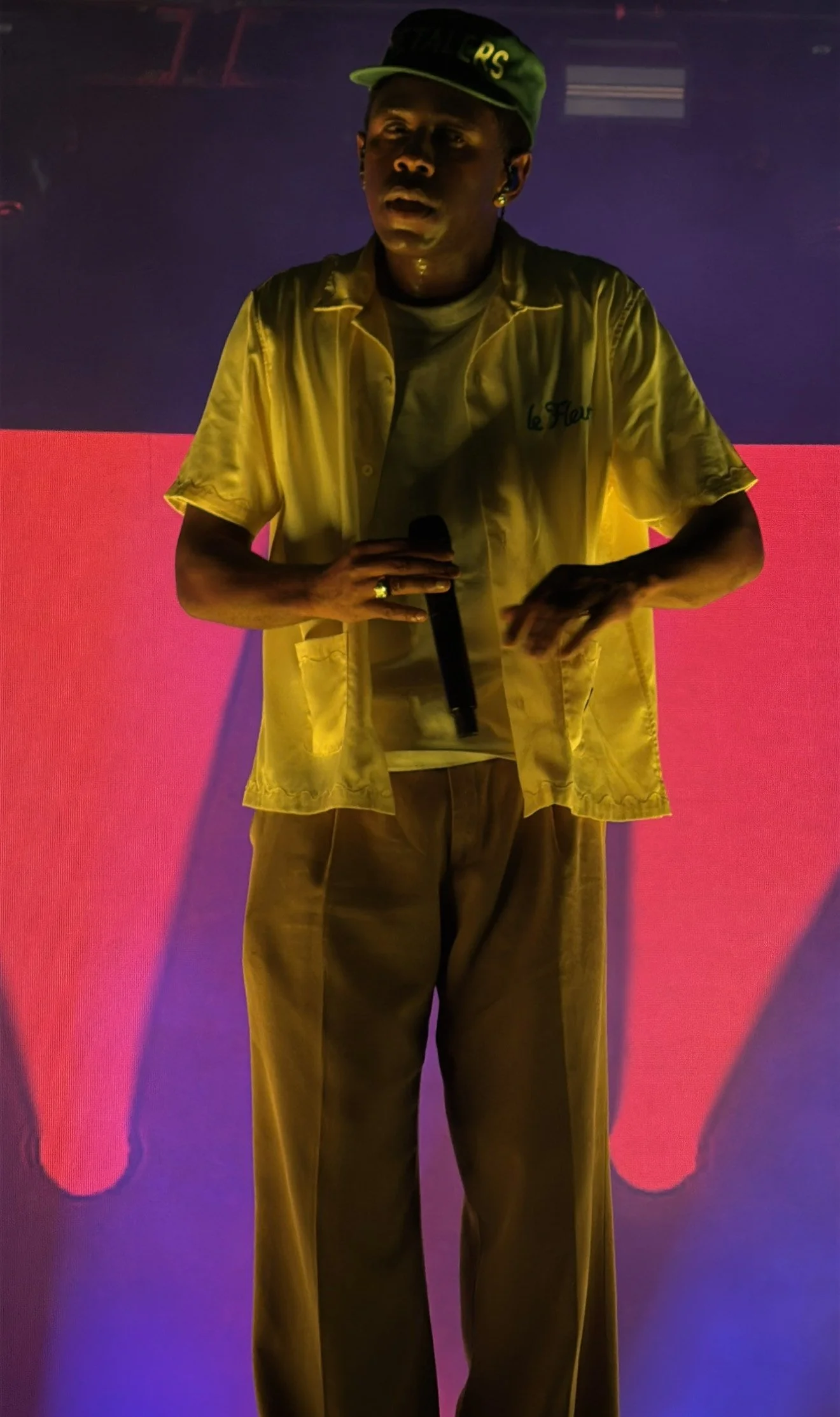 A person wearing a yellow shirt and beige pants, holding a microphone, standing on a stage with pink and purple lighting.