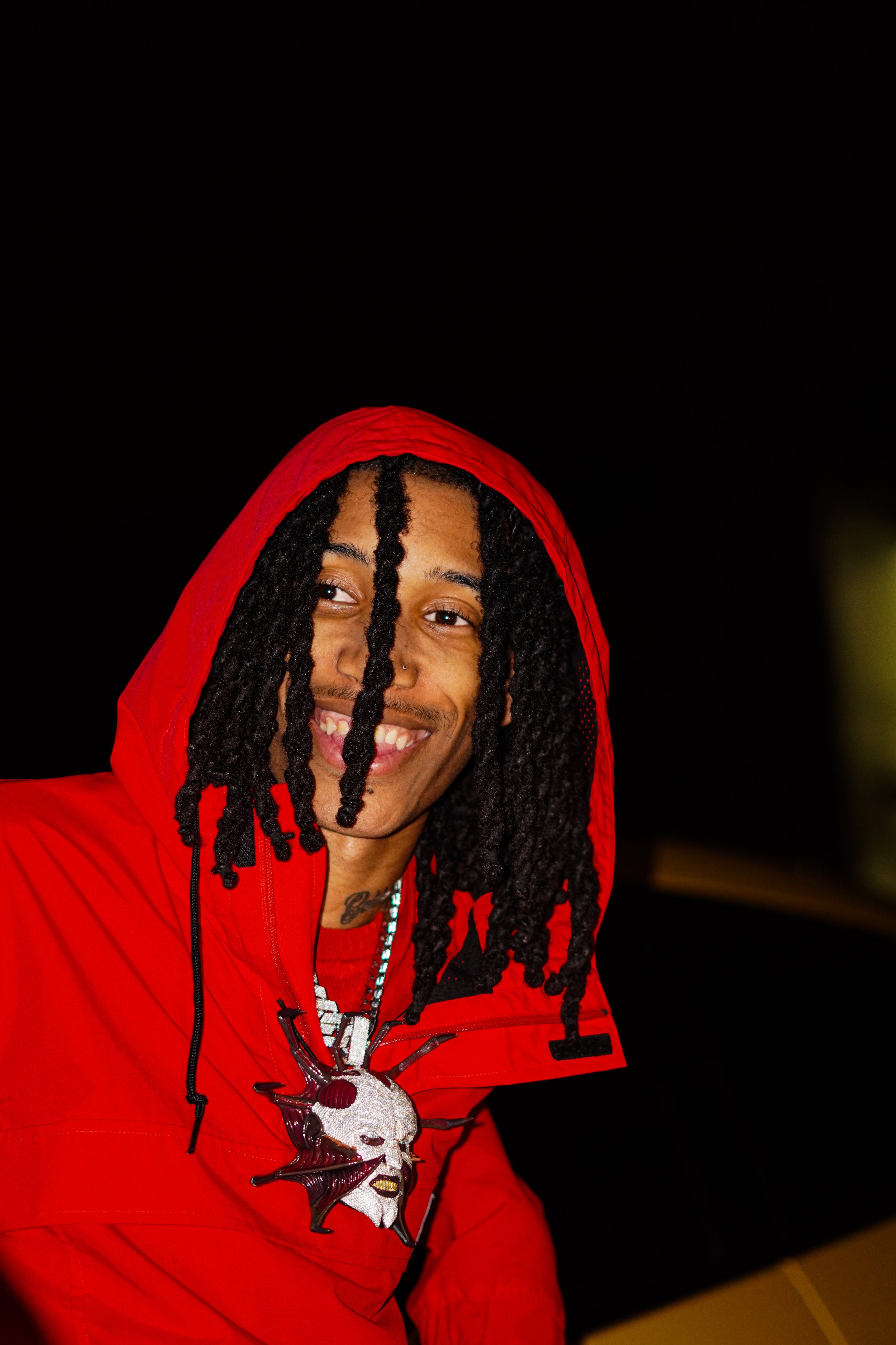 A smiling man with dreadlocks wearing a red hoodie and a large, distinctive skull pendant necklace.