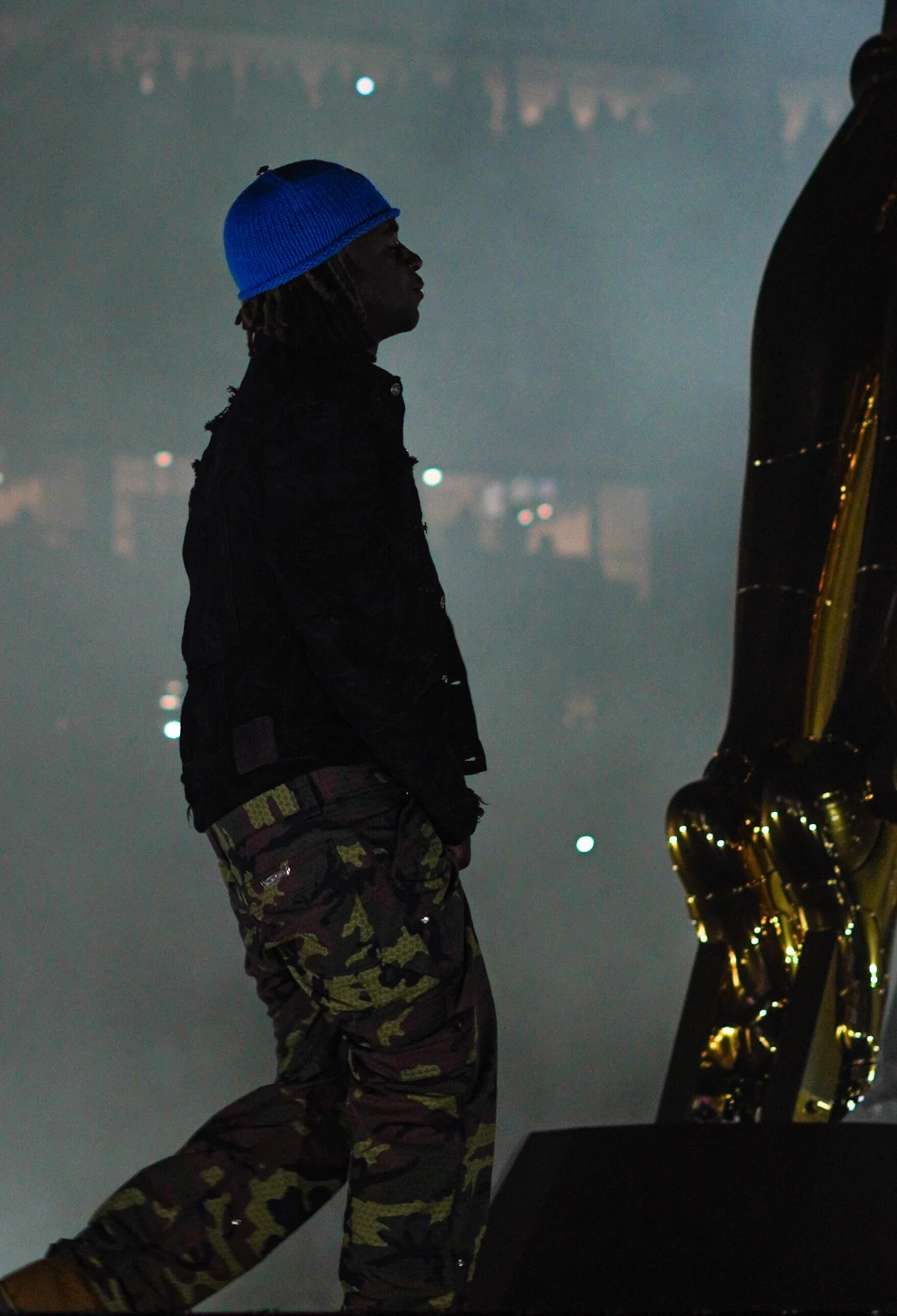 A silhouette of a man wearing a blue beanie, black jacket, and camouflage pants, standing beside a golden saxophone with a foggy, cityscape background at night.