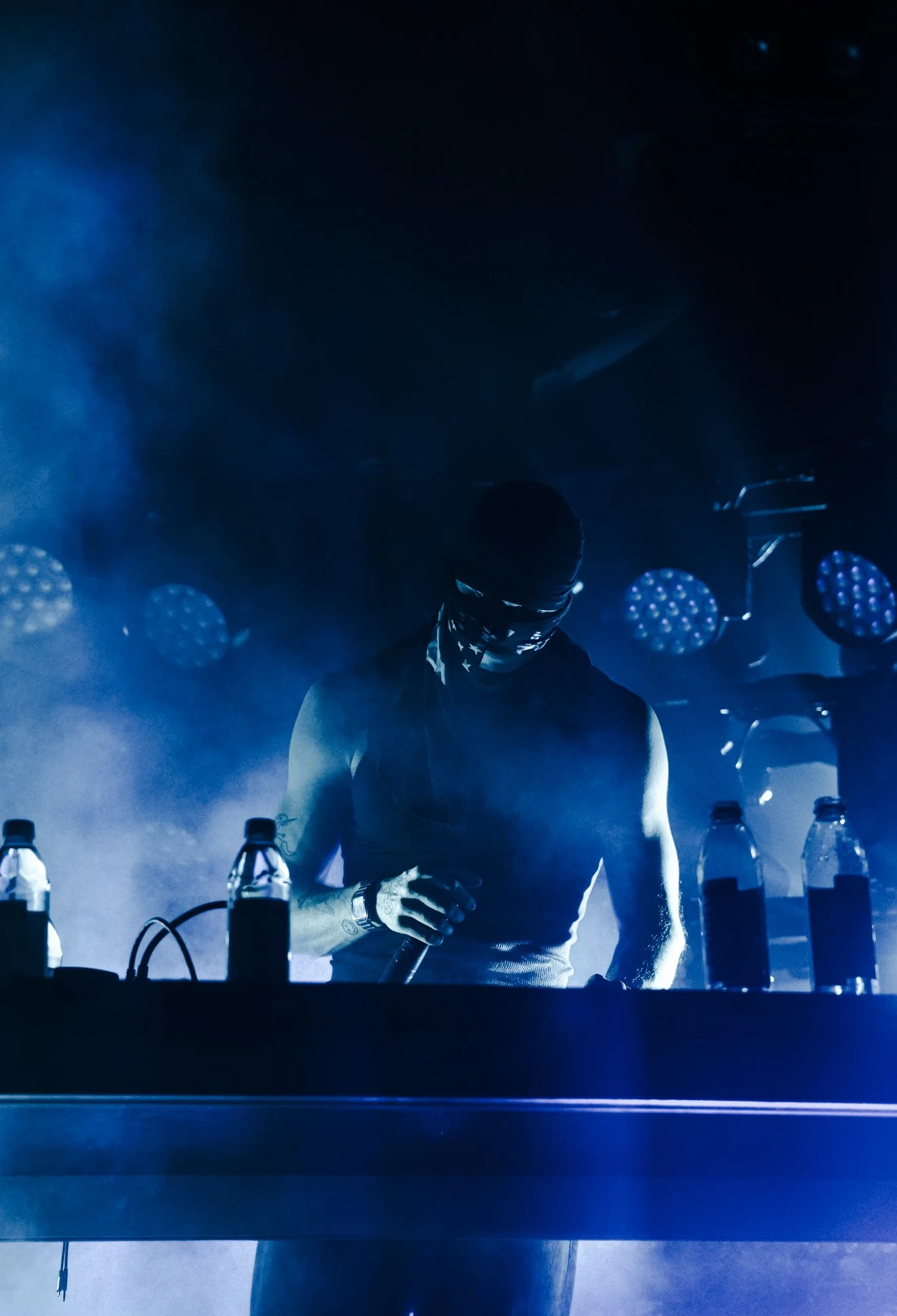A DJ wearing a mask and sleeveless shirt performing at a dark concert with blue lighting and water bottles on the DJ booth.