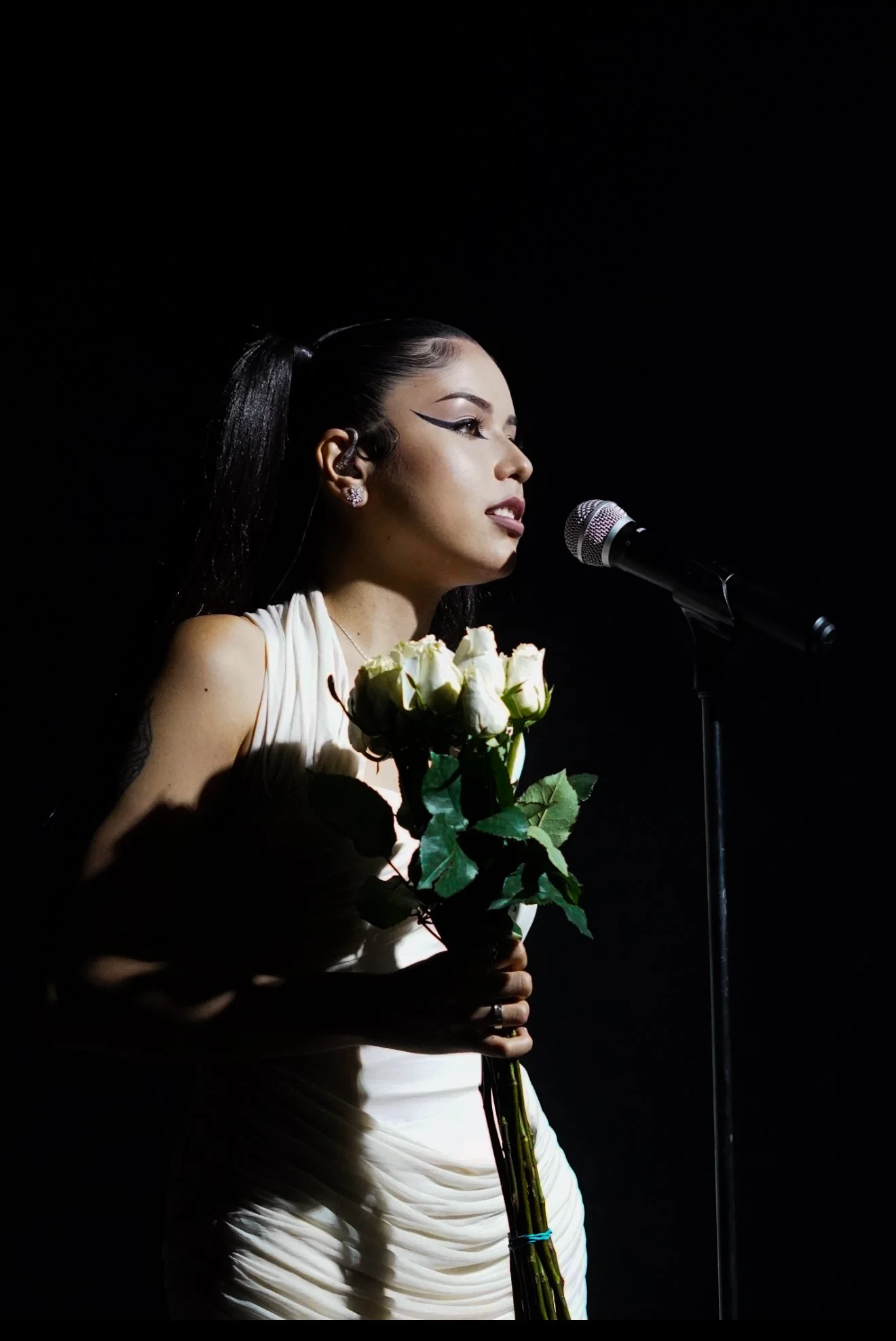 A woman with long dark hair and winged eyeliner stands in front of a microphone, holding a bouquet of white roses, with a dark background.