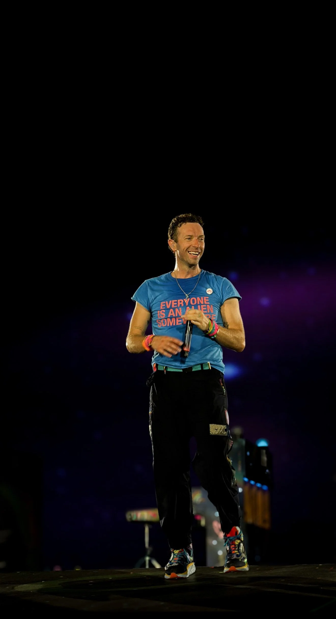A man with short curly hair, wearing a blue shirt with the text "Everyone is an alien somewhere," black pants, and colorful sneakers, is smiling and holding a microphone on a stage with a dark background.