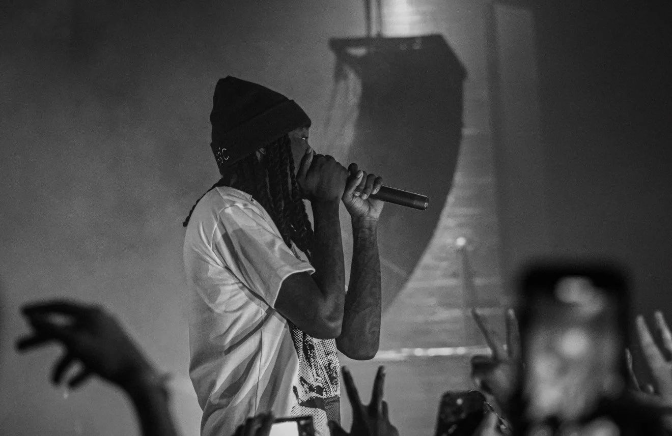 A person with dreadlocks, wearing a beanie and a T-shirt, holding a microphone and performing on stage in black and white, with an audience taking photos and videos.