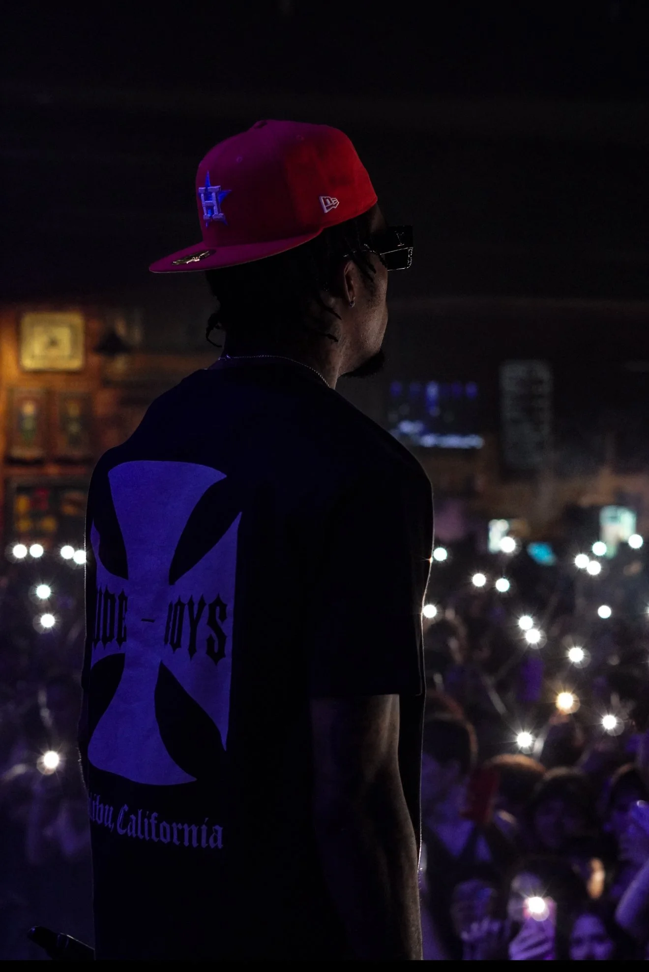 A man wearing a red baseball cap, black sunglasses, and a black T-shirt with a large design on the back stands on stage, illuminated by his phone lights, in front of a crowd in a dark bar or club.