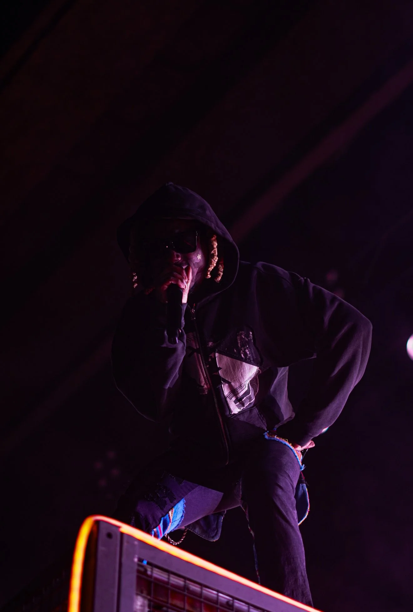 A person dressed in dark clothing, wearing a hoodie and sunglasses, holding a microphone, performing on stage with purple lighting.