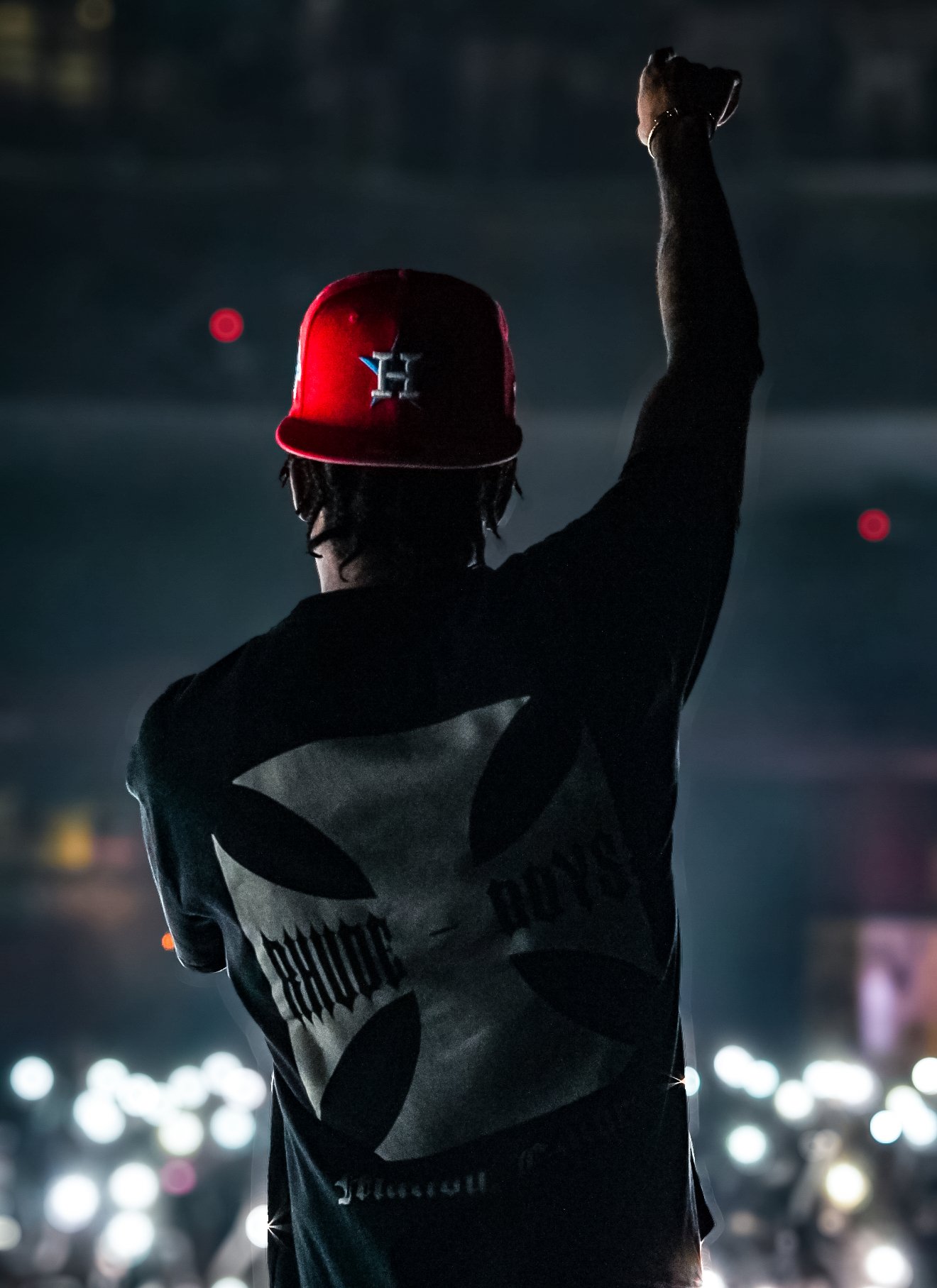 A rapper on stage with a dark background, wearing a red cap and a black long-sleeved shirt with a skull design and the word 'BOUNTY' on the back, raising one arm.