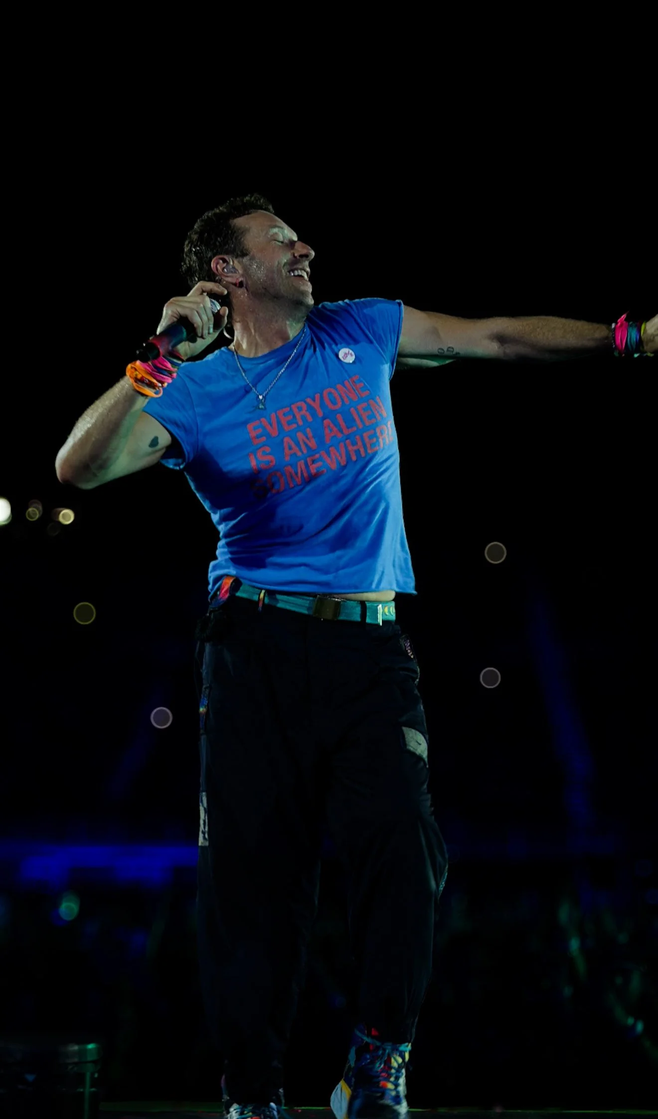 A man performing on stage at night, holding a microphone, wearing a blue t-shirt with the text "Everyone is an alien somewhere", black pants, colorful wristbands, and sneakers, with a dark background and some stage lights.