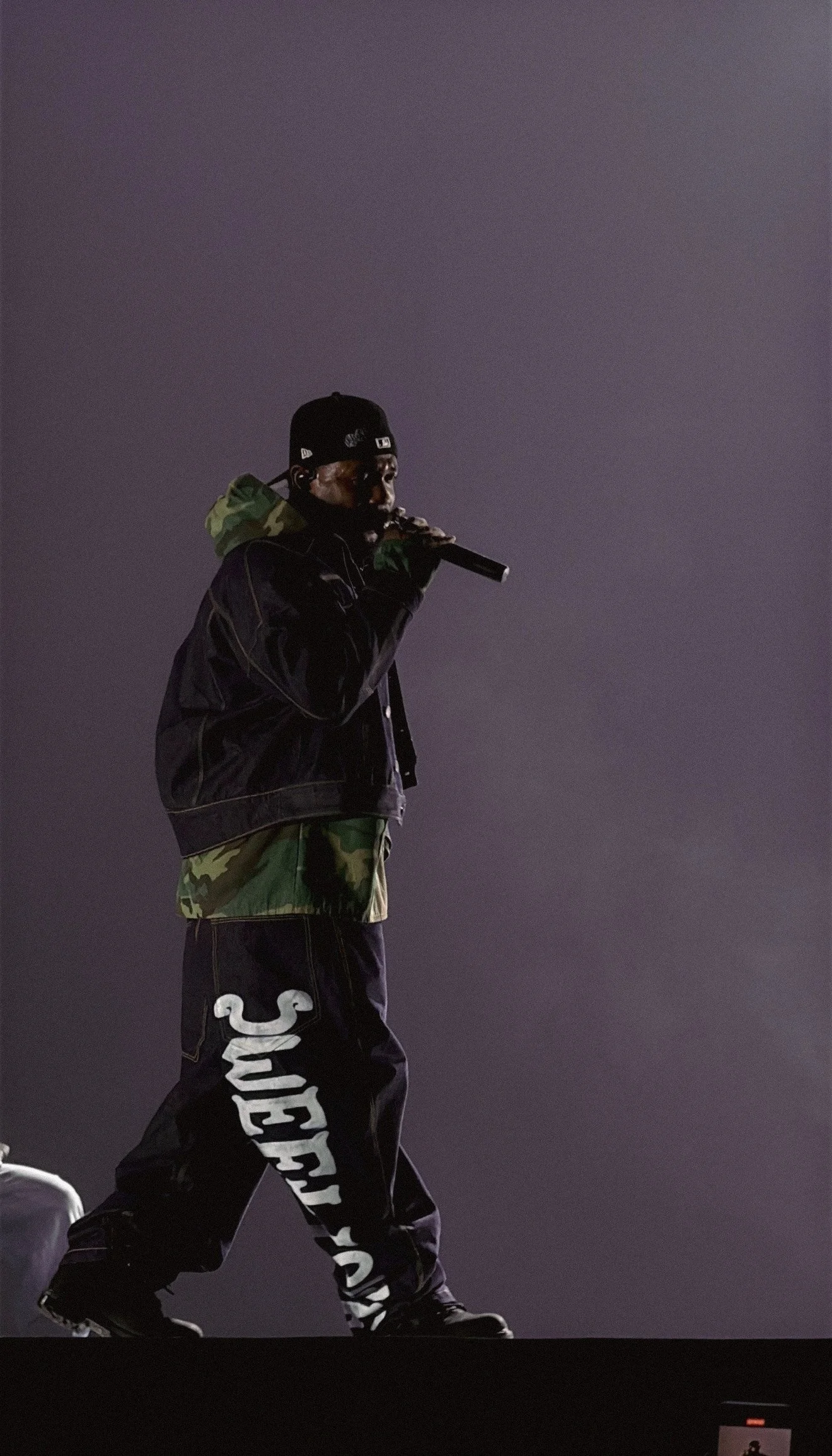 A person wearing a black cap and jacket, with green camouflage shirt and pants, holding a microphone and standing against a plain dark background.