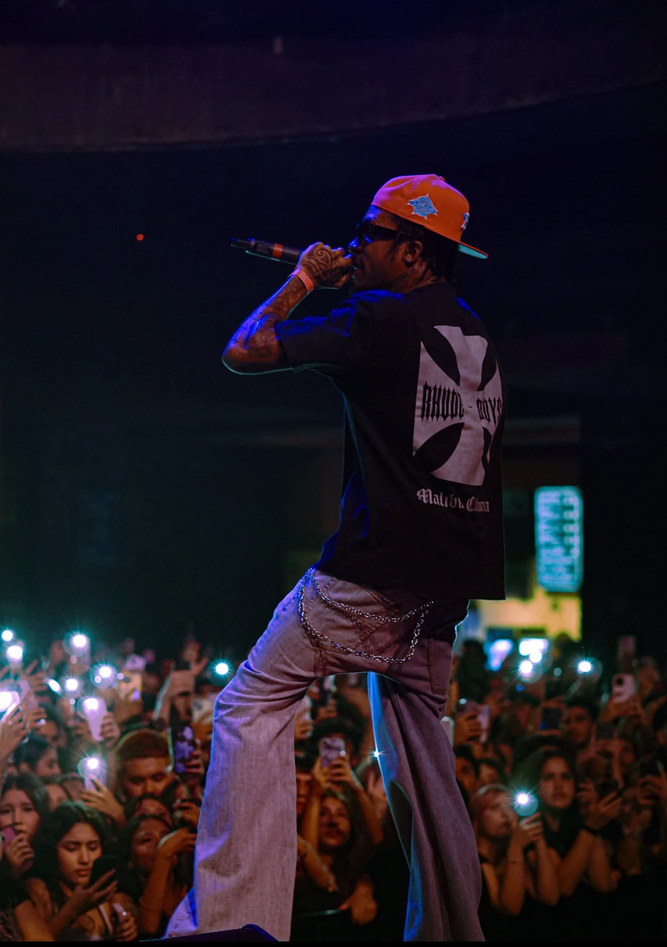 A performer wearing a black Rhude Rude t-shirt, beige baggy pants with chains, a backward orange cap, sunglasses, and tattoos, holds a microphone and performs on stage in front of a crowd with many holding up smartphones.