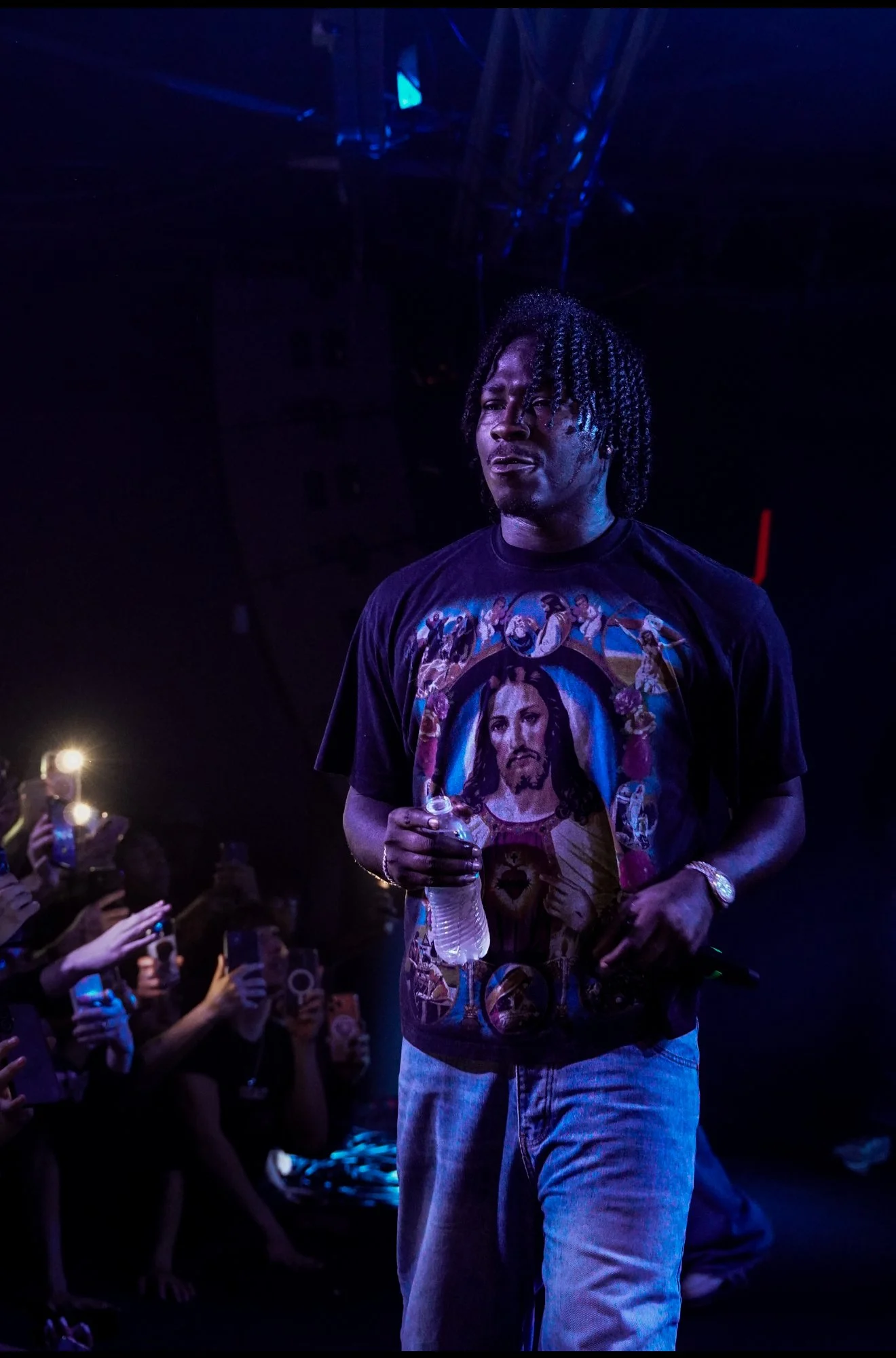 A man with dreadlocks wearing a Jesus-themed T-shirt walking through a crowd at a concert or event, holding a water bottle, with many people around taking photos.