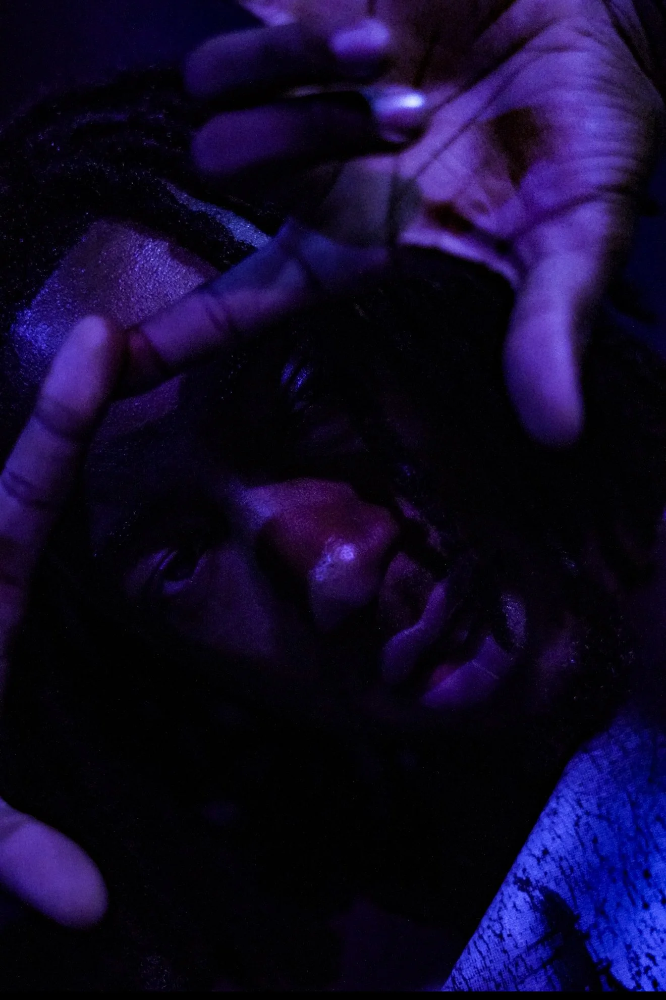 A person with dark skin and curly hair is lying down in a dark setting with purple lighting, with hands framing their face.