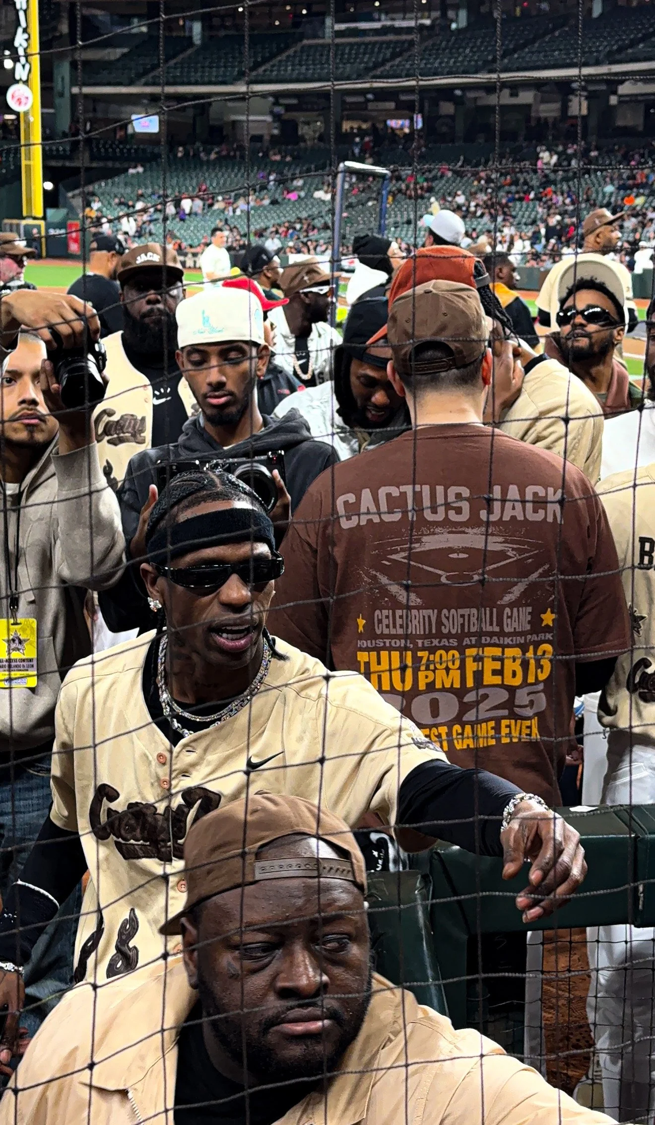 People gathered at a baseball game, viewed through a protective net. Some are taking pictures or videos, and the stadium is visible in the background.