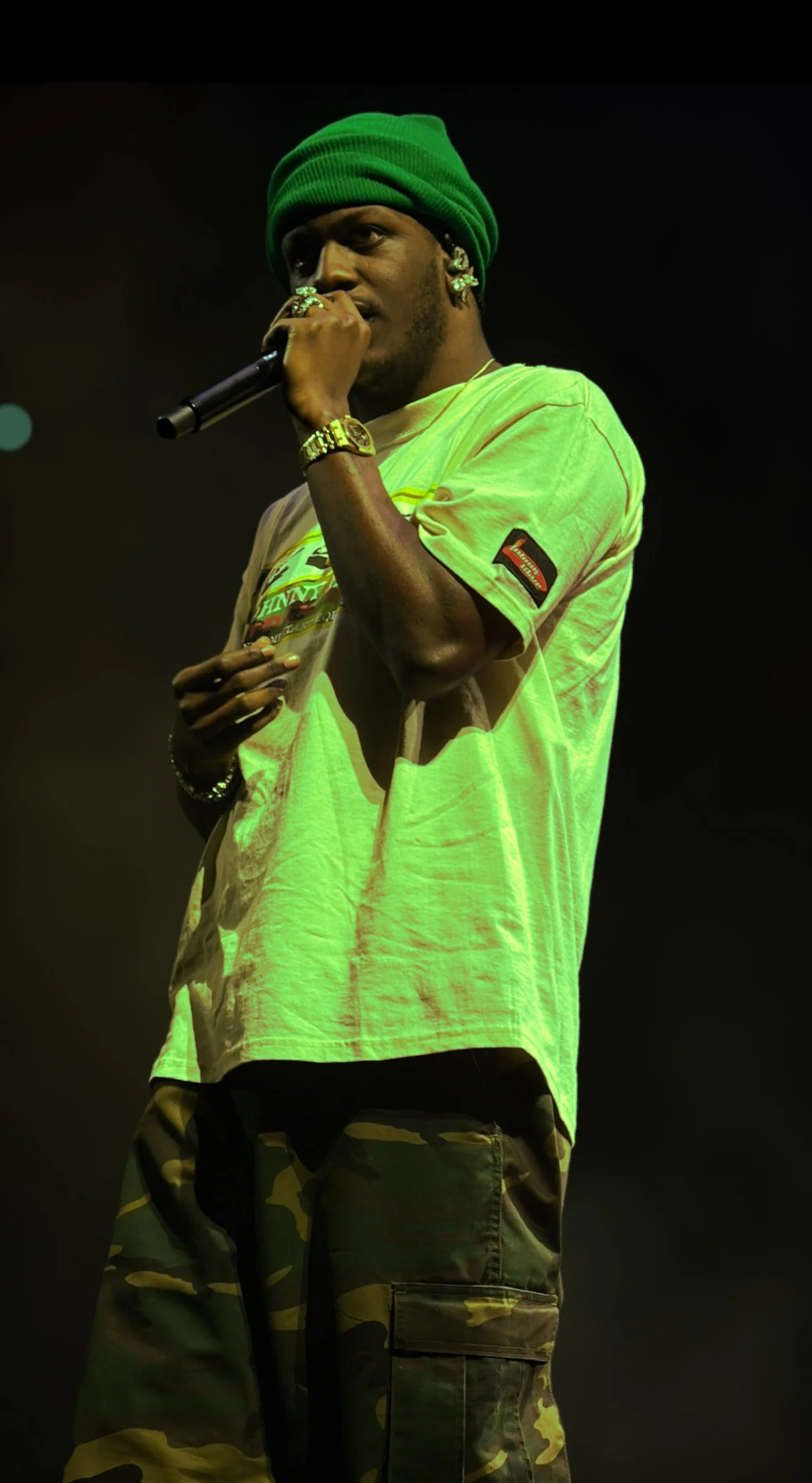 A young man wearing a green beanie, yellow graphic t-shirt, and camouflage cargo pants holding a microphone on stage.