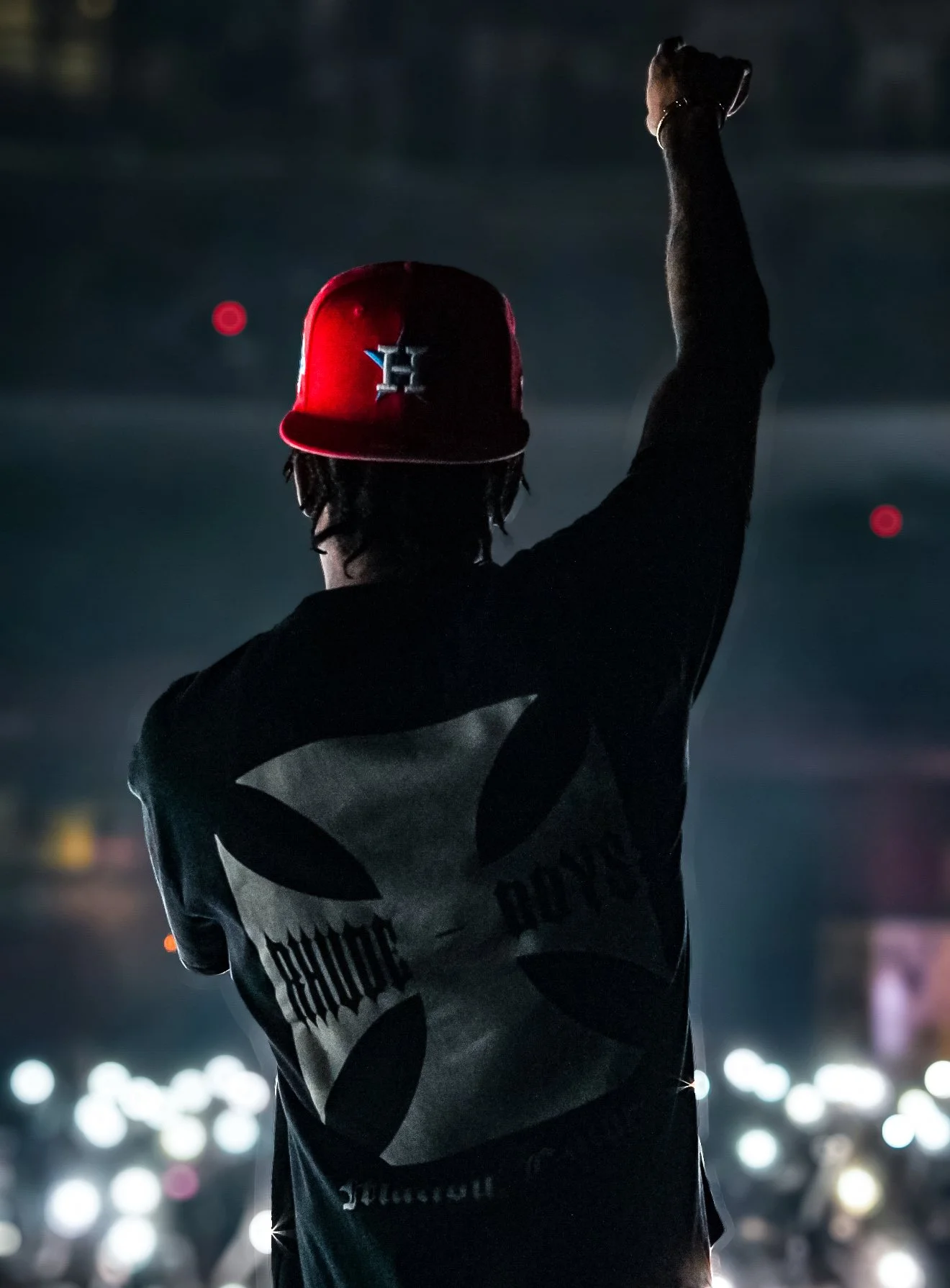 A person wearing a red baseball cap with a logo, a black jacket with a large graphic, standing with one arm raised at a nighttime event with bright lights in the background.