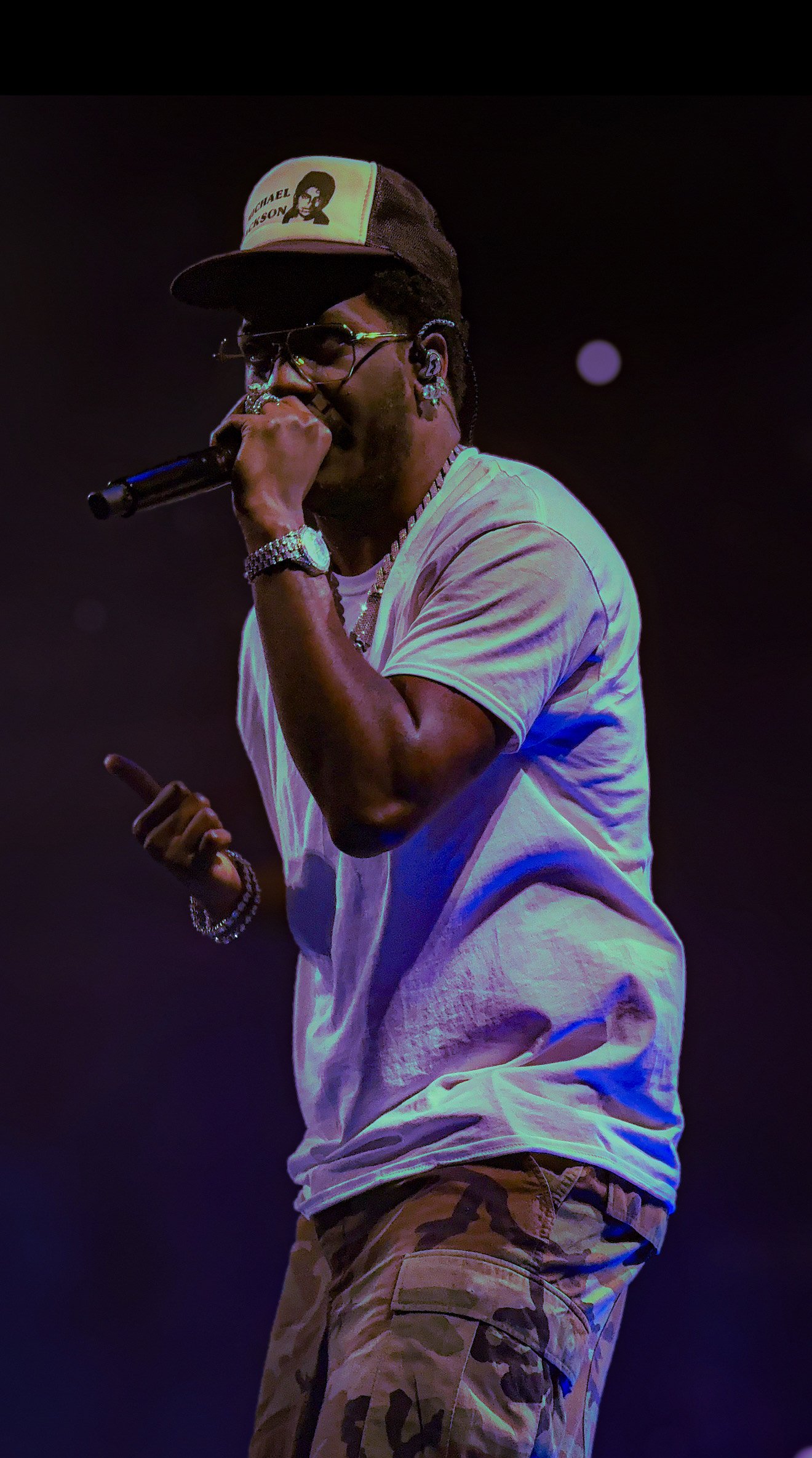 A male performer singing into a microphone on stage, wearing sunglasses, a cap with a graphic and text, a white t-shirt, camouflage pants, and jewelry, with a dark background and stage lighting.