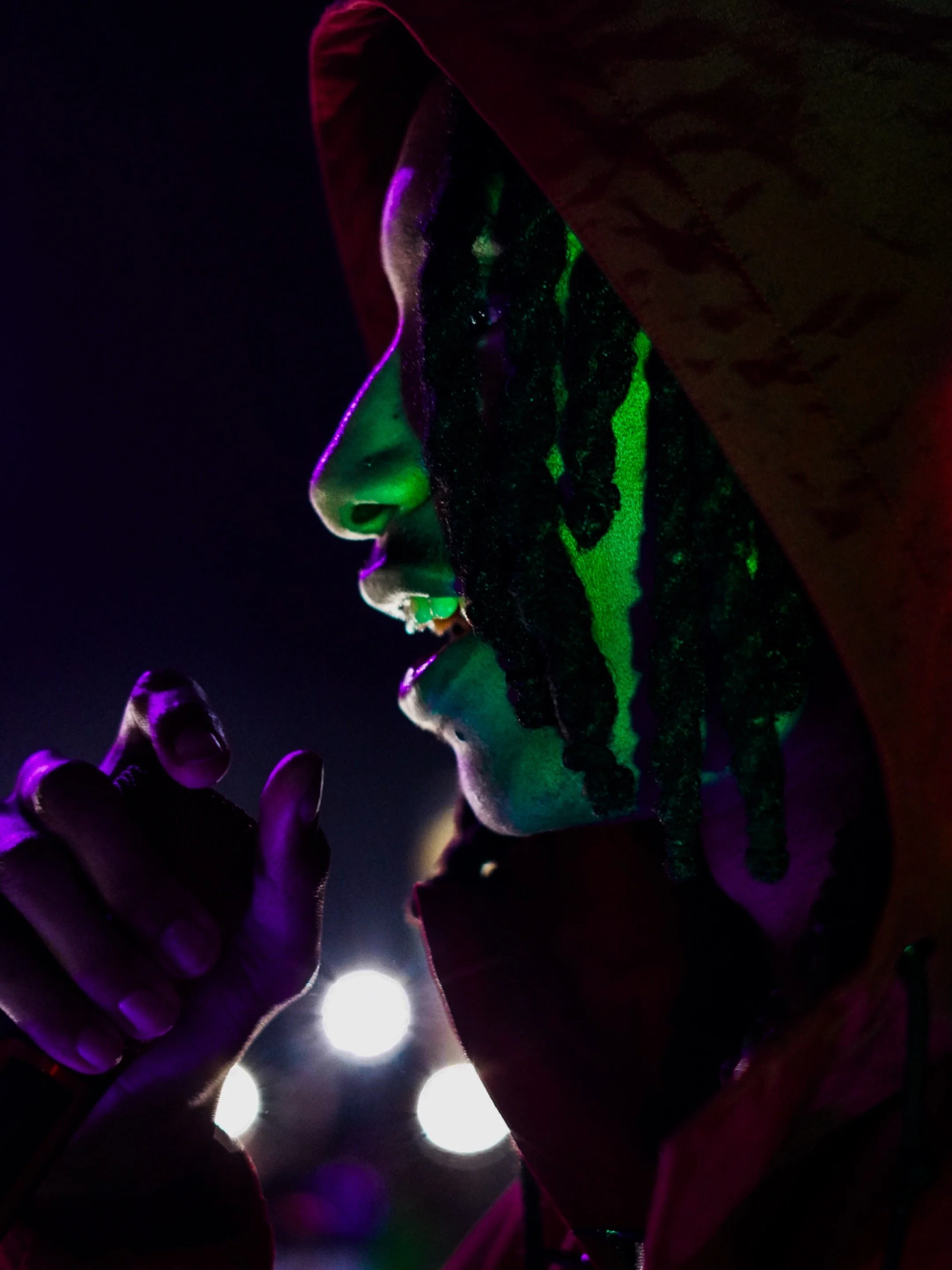 A person wearing a hooded jacket with the hood up, illuminated by colorful and neon lighting, appears to be smoking or holding a cigarette close to their face, with a dark background and bright lights behind them.