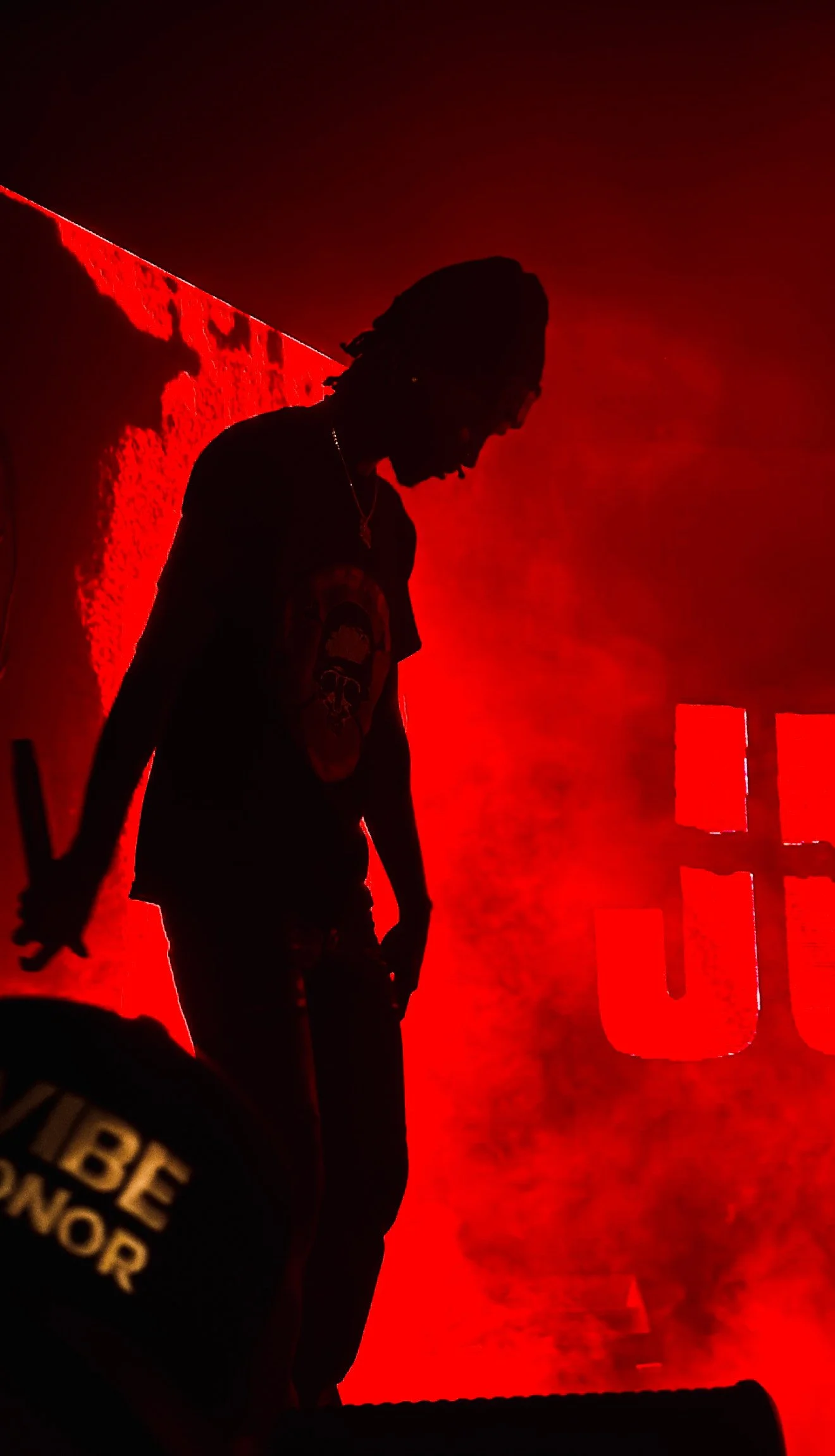 Silhouette of a person with dreadlocks in front of a bright red illuminated background, holding a stick or bat, with part of the word 'U' visible.