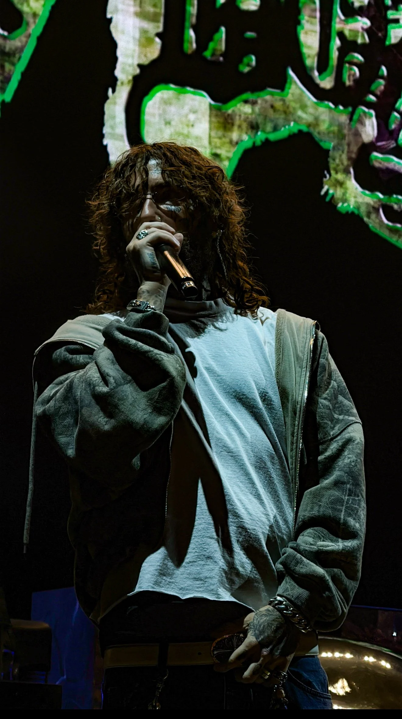 A man with long curly hair, wearing glasses, a white t-shirt, and a gray jacket, holding a microphone on stage in a dark setting with a colorful background.