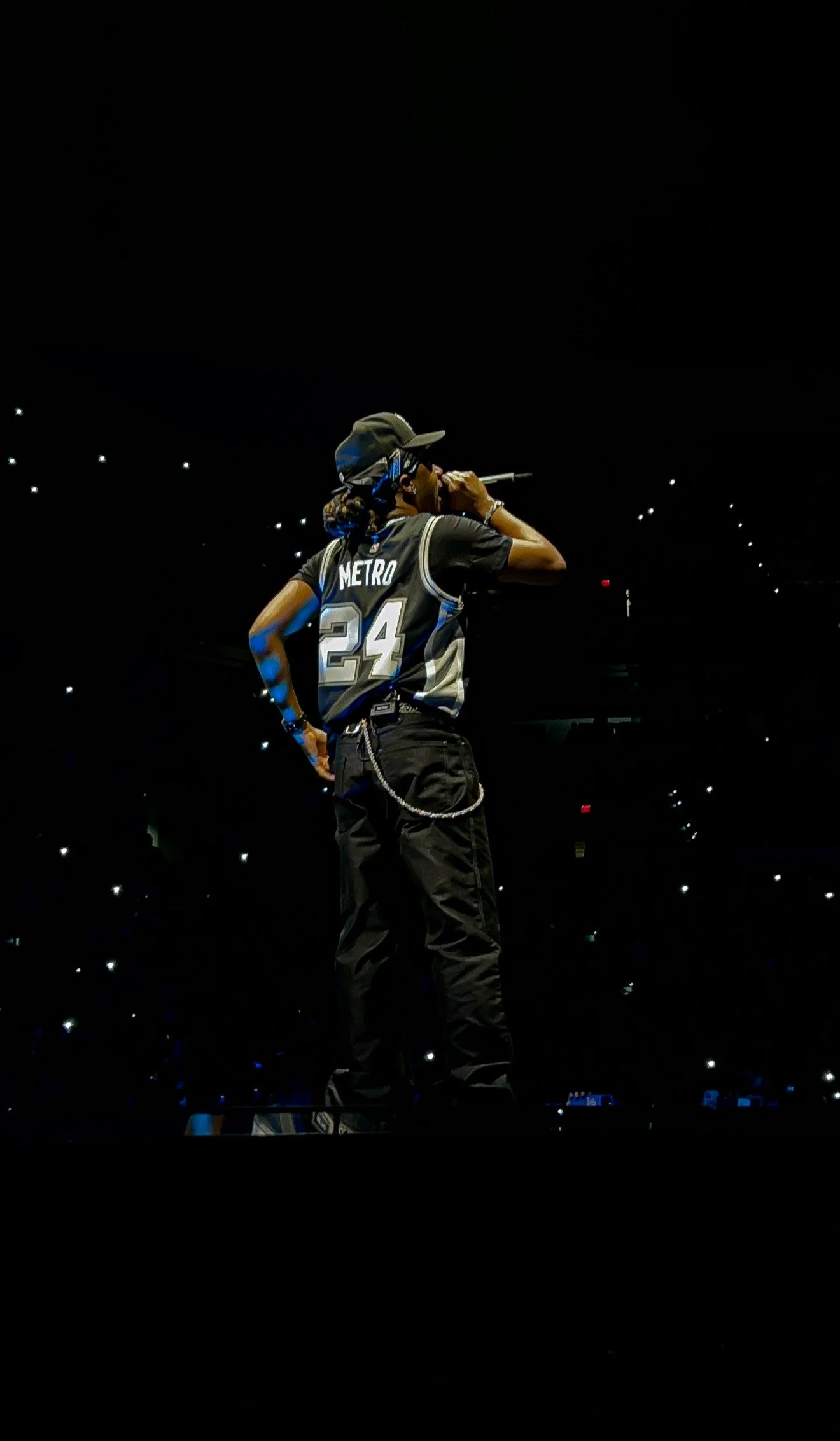 Performer on stage singing into a microphone, wearing a sports jersey with 'METRO' and the number 24, in a dark setting with small lights.