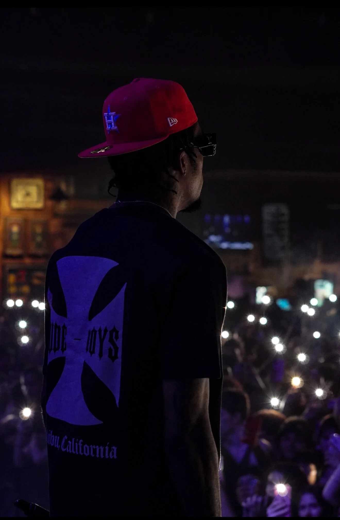 Person wearing a red cap and black t-shirt with a cross logo, performing on stage in front of a crowd at night.