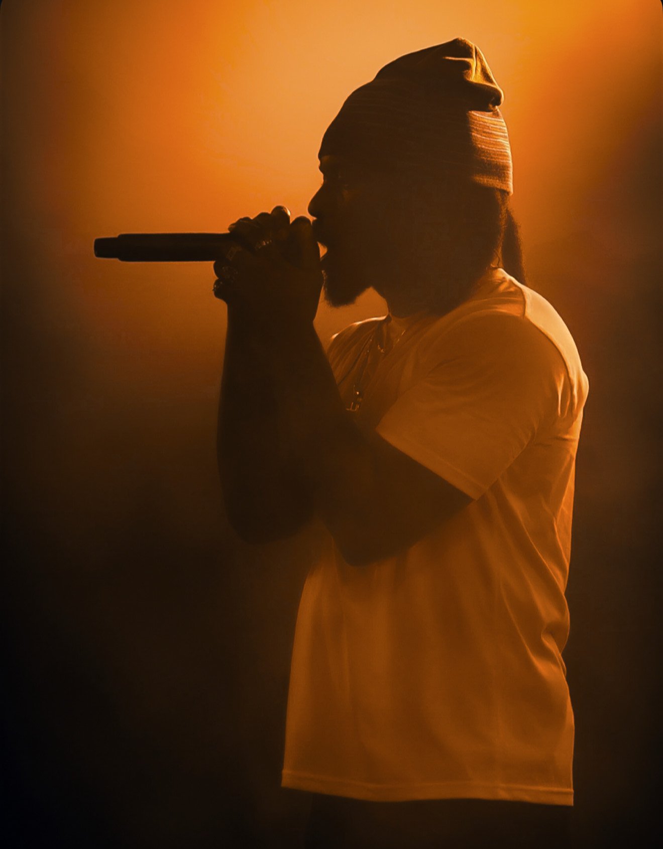 Silhouette of a man with dreadlocks and a beard singing into a microphone, illuminated by warm orange stage lighting, wearing a headscarf and a yellow shirt.