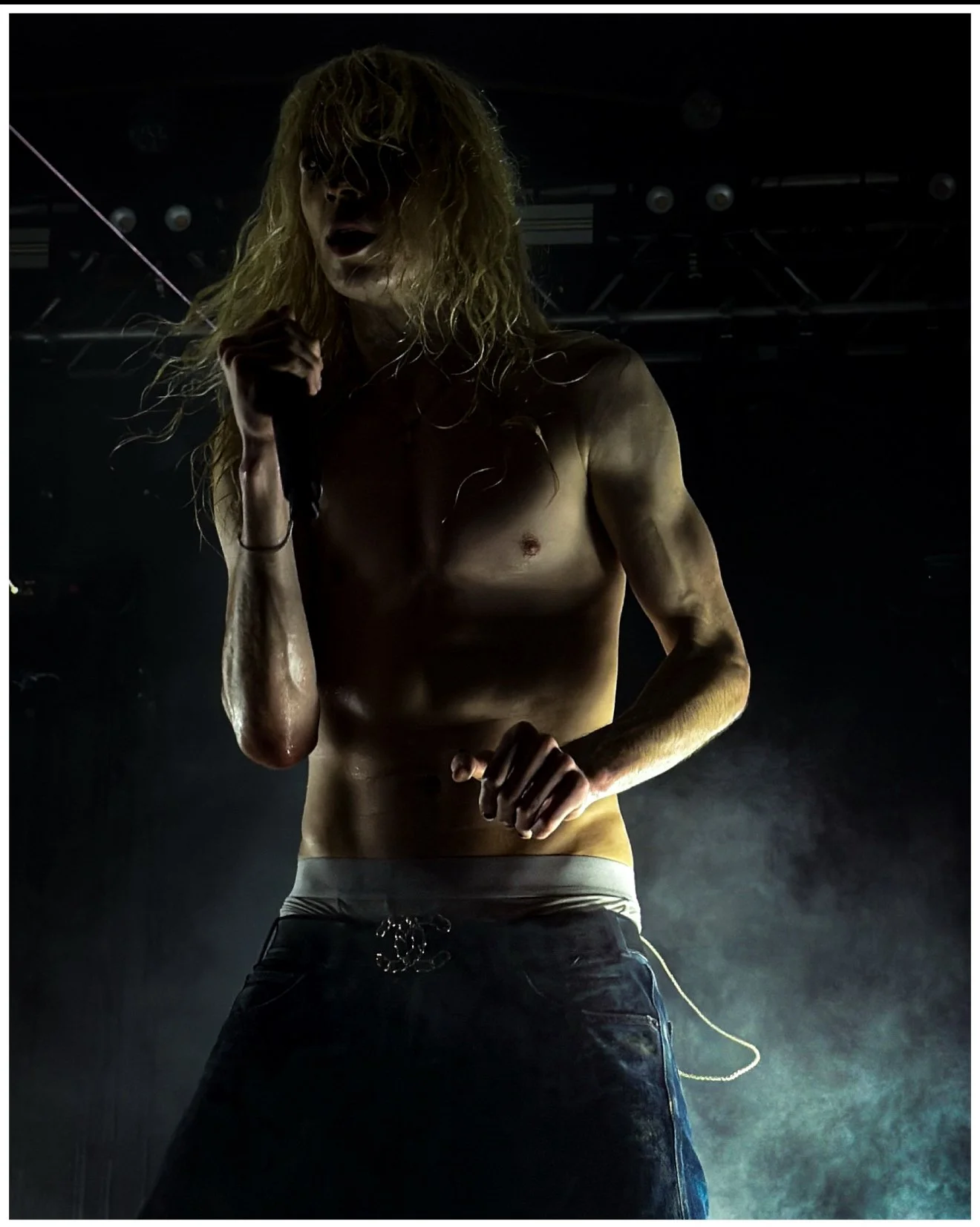 A shirtless male performer with long, curly blond hair, holding a microphone and standing on stage with fog and stage lights in the background.